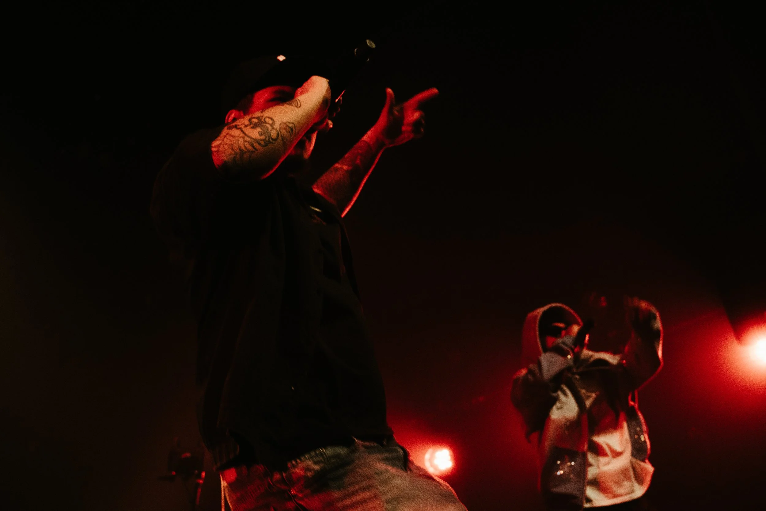 Deux rappeurs sur scène, un en t-shirt noir avec des tatouages et une casquette, l'autre en hoodie avec le capuchon sur la tête, sous un éclairage rouge.