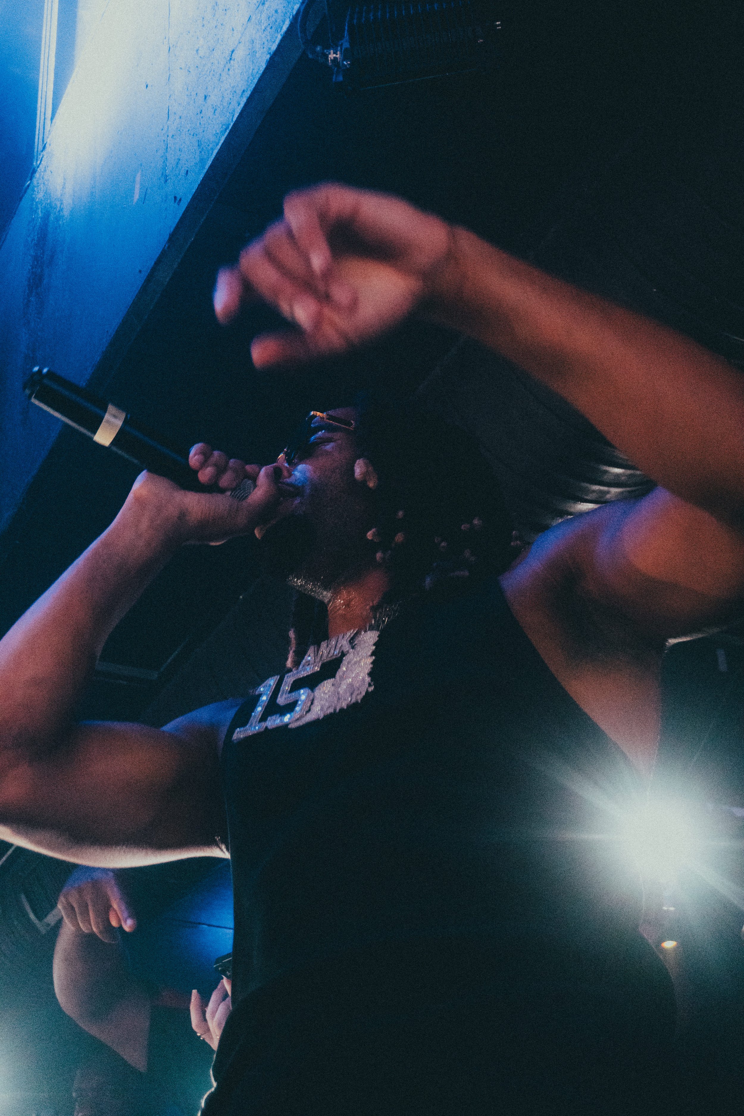 Une femme avec des dreadlocks, portant un t-shirt noir avec le numéro 15, chante dans un microphone dans un environnement sombre avec des lumières bleues et un strong rétro.