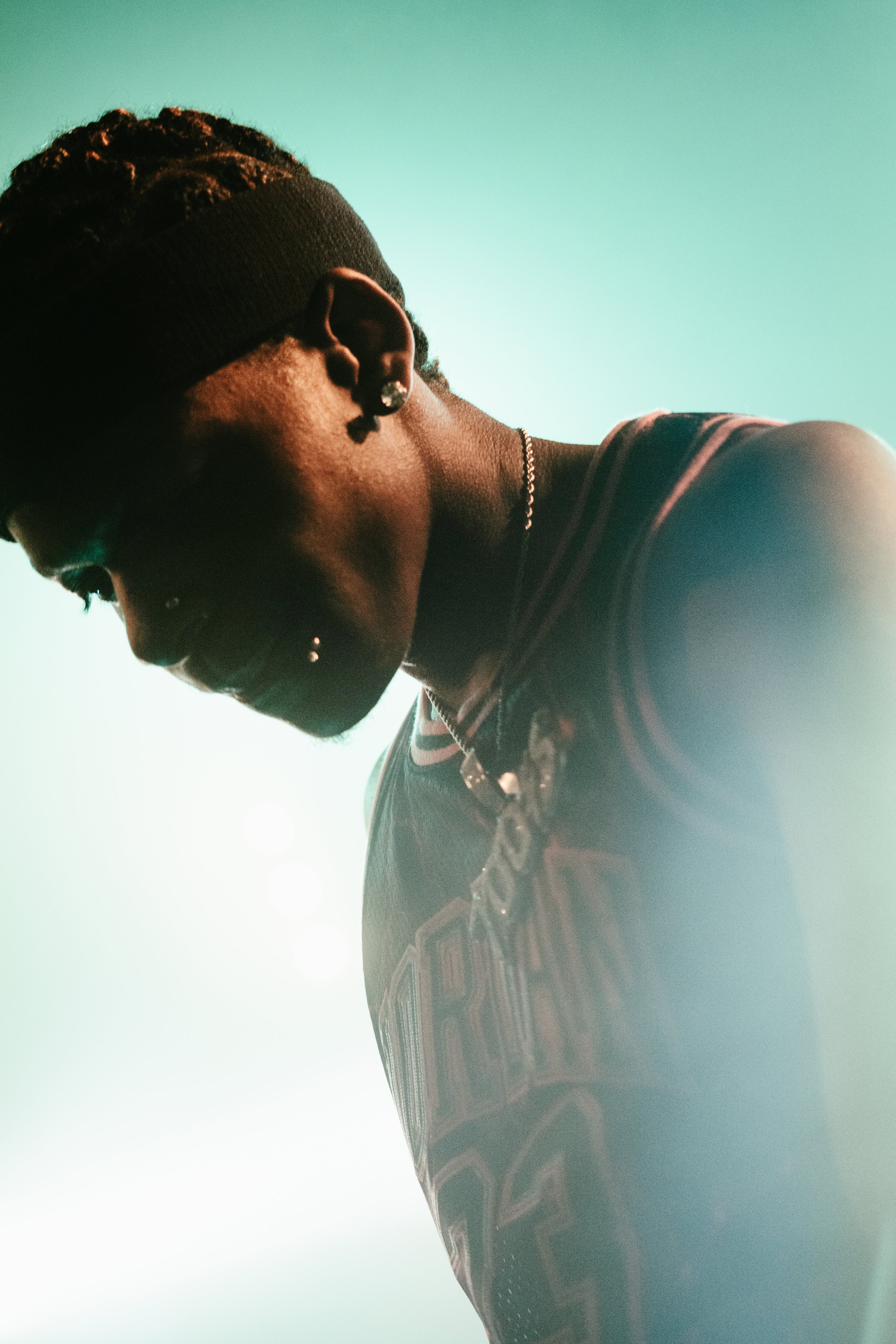 Jeune homme portant un bandeau noir, une robe à rayures et des bijoux, regardant vers le bas dans une lumière douce.