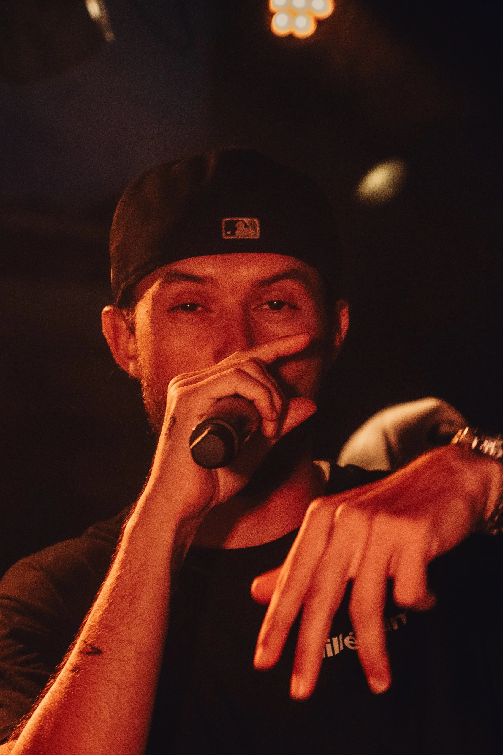 Une personne portant une casquette de baseball noire avec le logo de la MLB, tenant un microphone, dans un environnement sombre avec des lumières chaudes, semblant chanter ou parler.