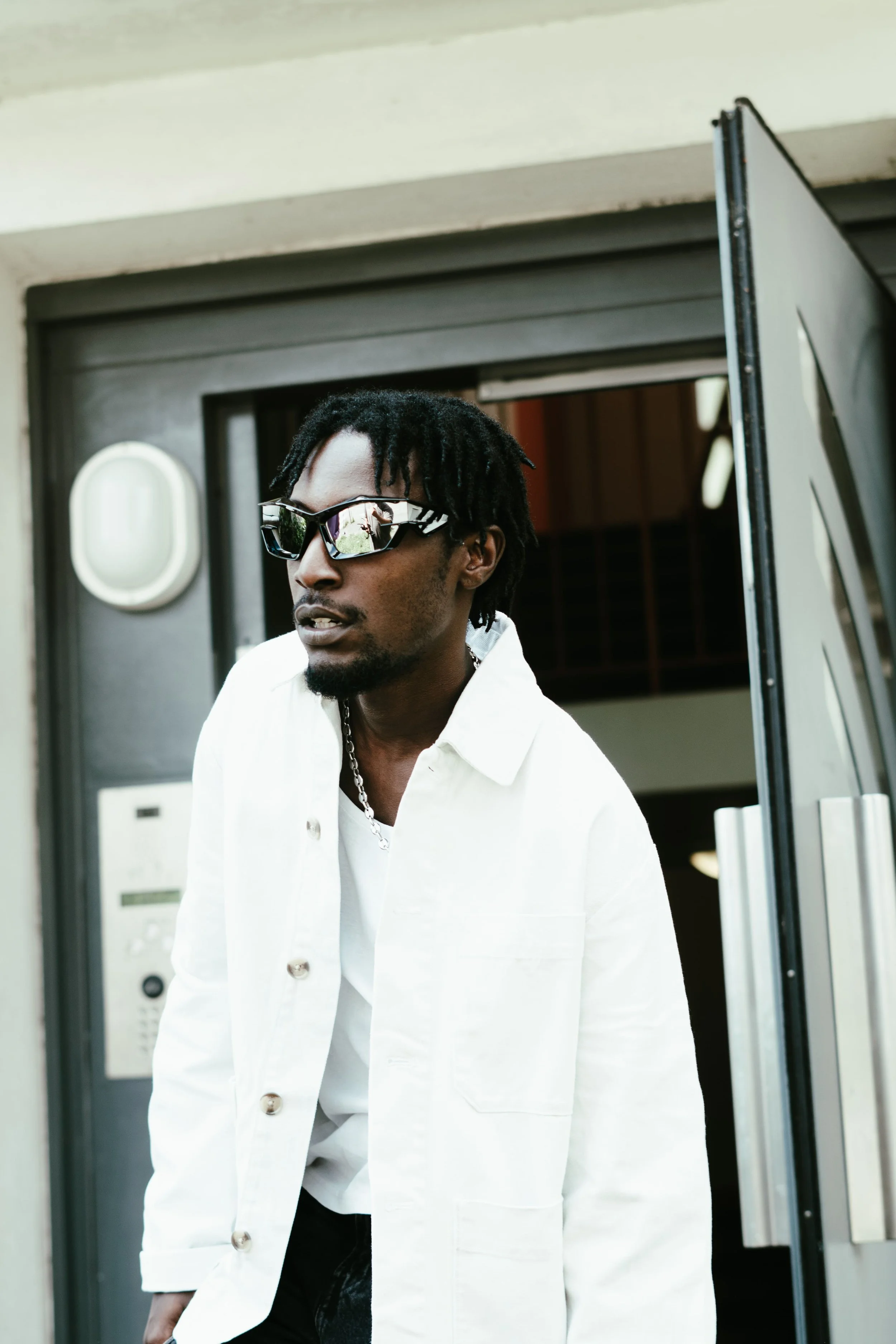 Jeune homme portant des lunettes de soleil, un manteau blanc, et une chaîne, sortant d'un bâtiment