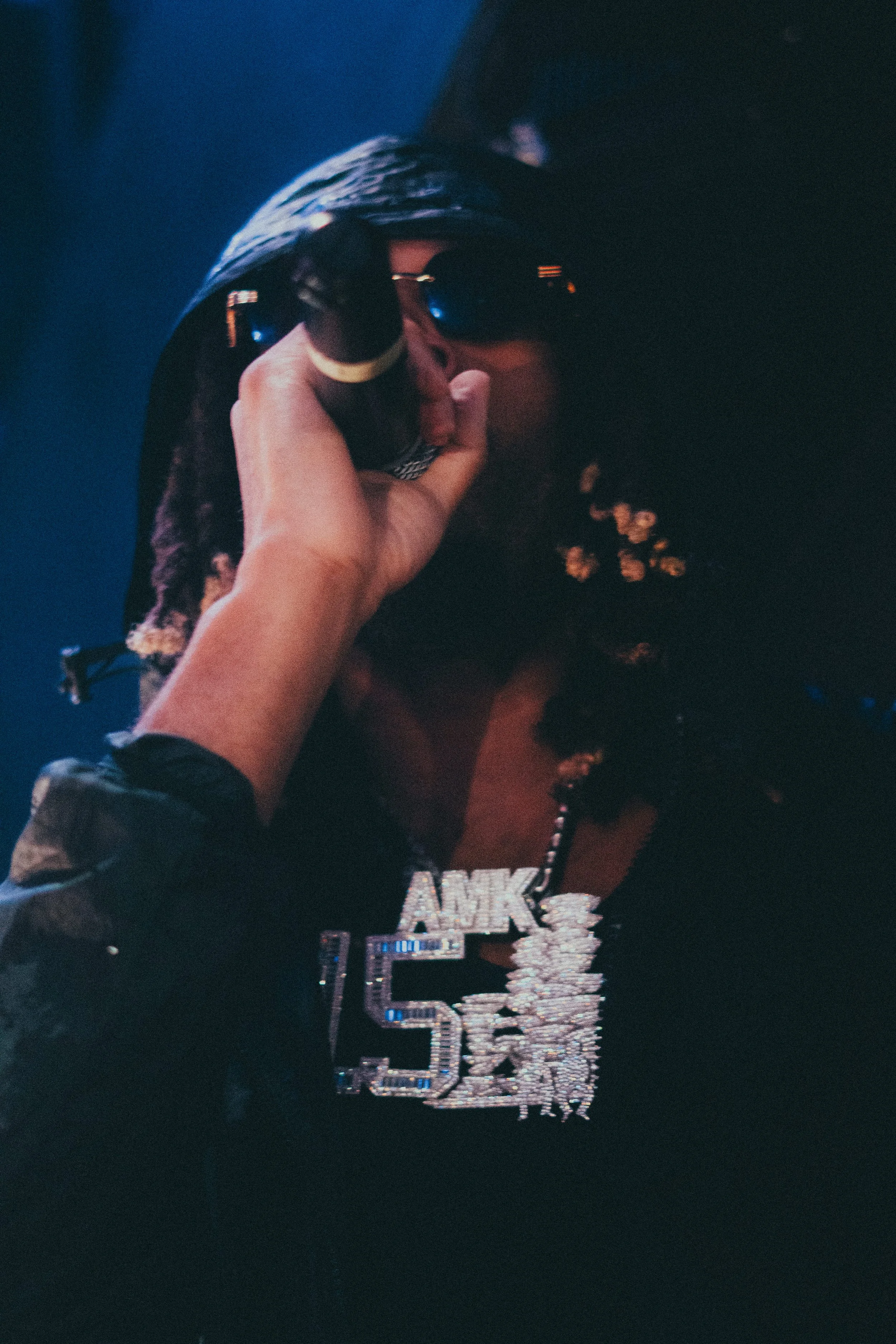 Personne portant des lunettes de soleil, un collier en chaîne brillante avec l'inscription 'BANK 150K', un hoodie noir, tenant un cigare.