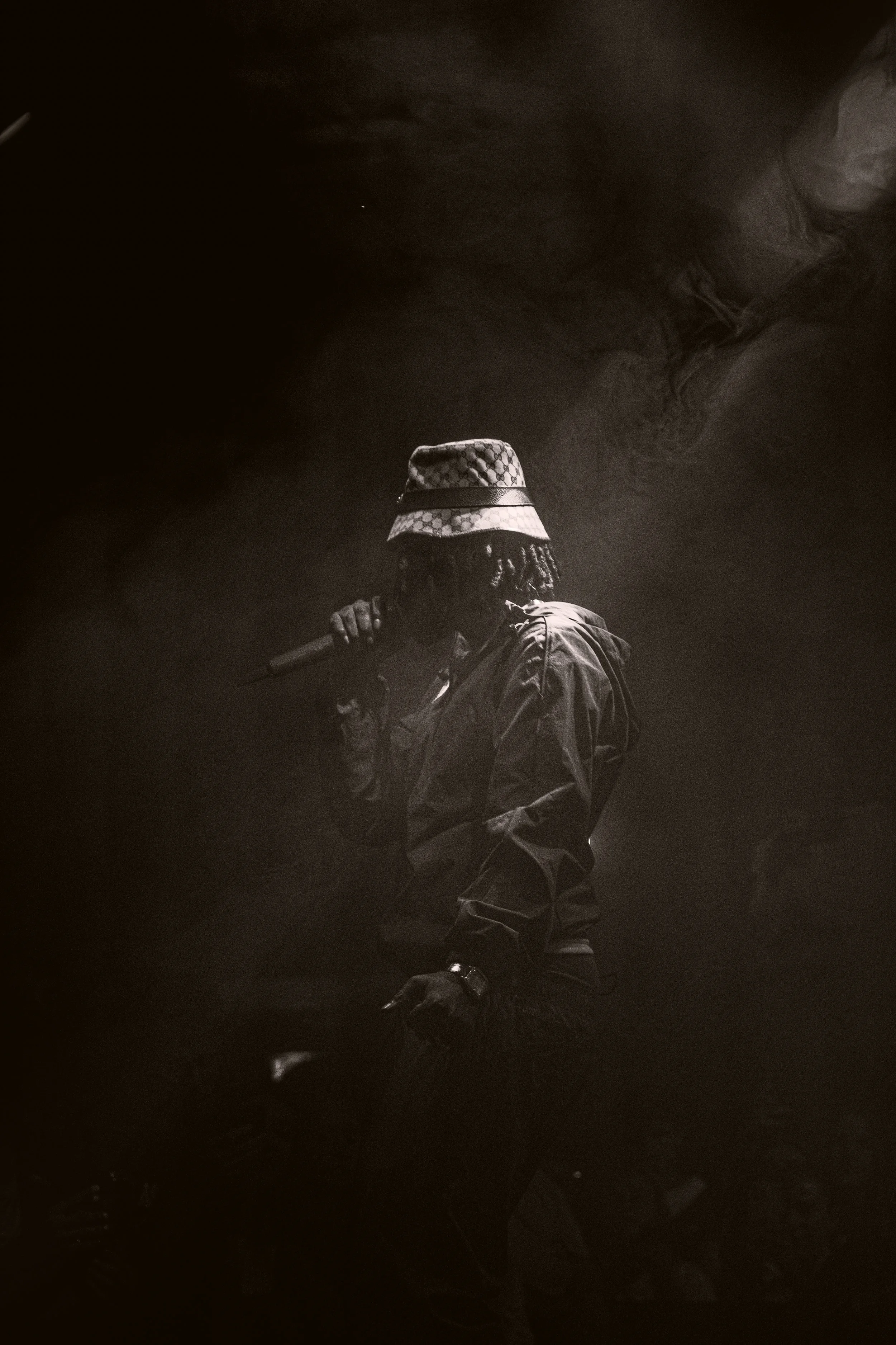 Une personne portant un chapeau à motifs et une veste sombre, tenant un microphone dans un environnement sombre et brumeux.
