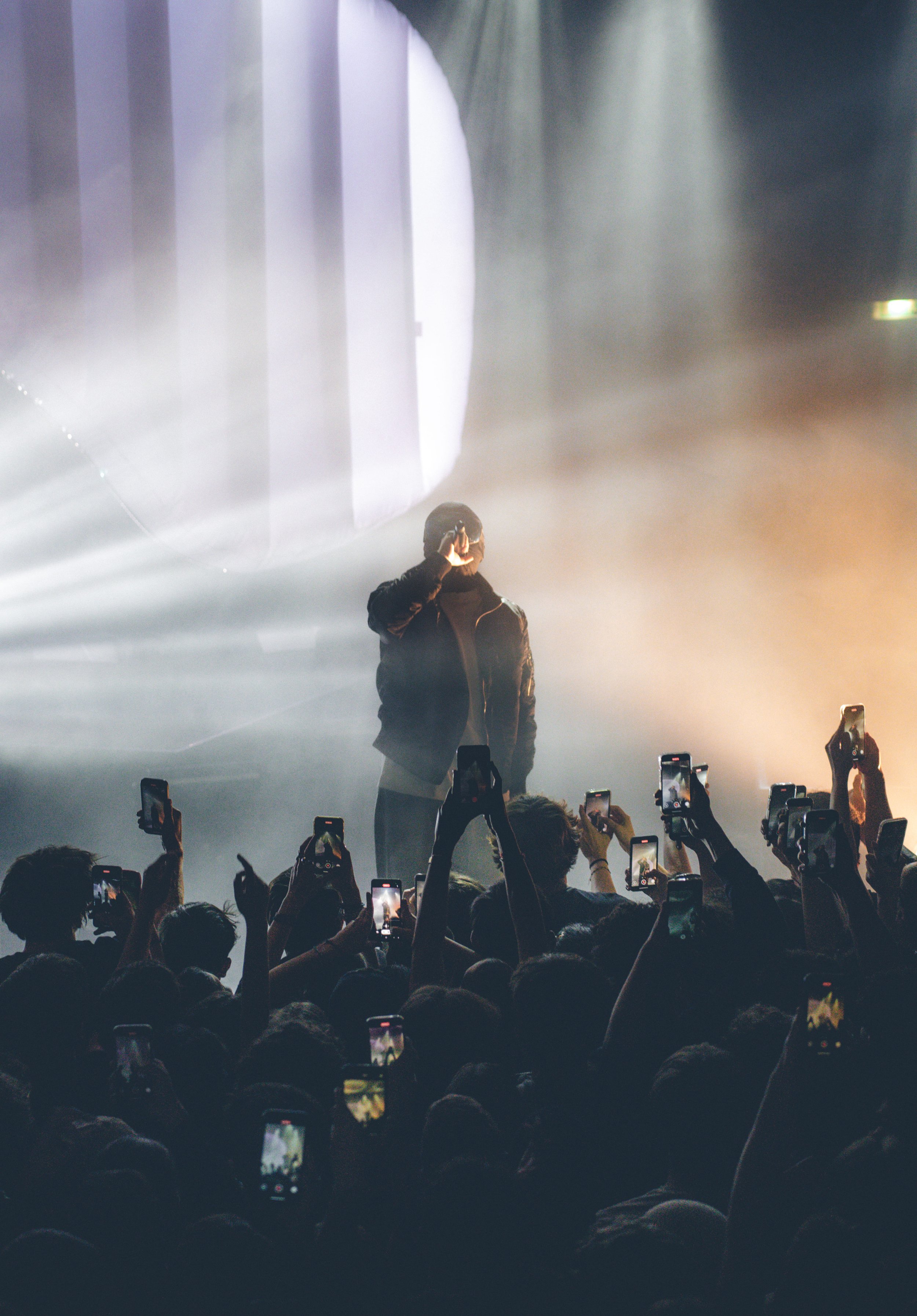 Un chanteur ou artiste sur scène lors d'un concert, entouré de fumée, avec un public en train de prendre des photos avec leurs téléphones portables.
