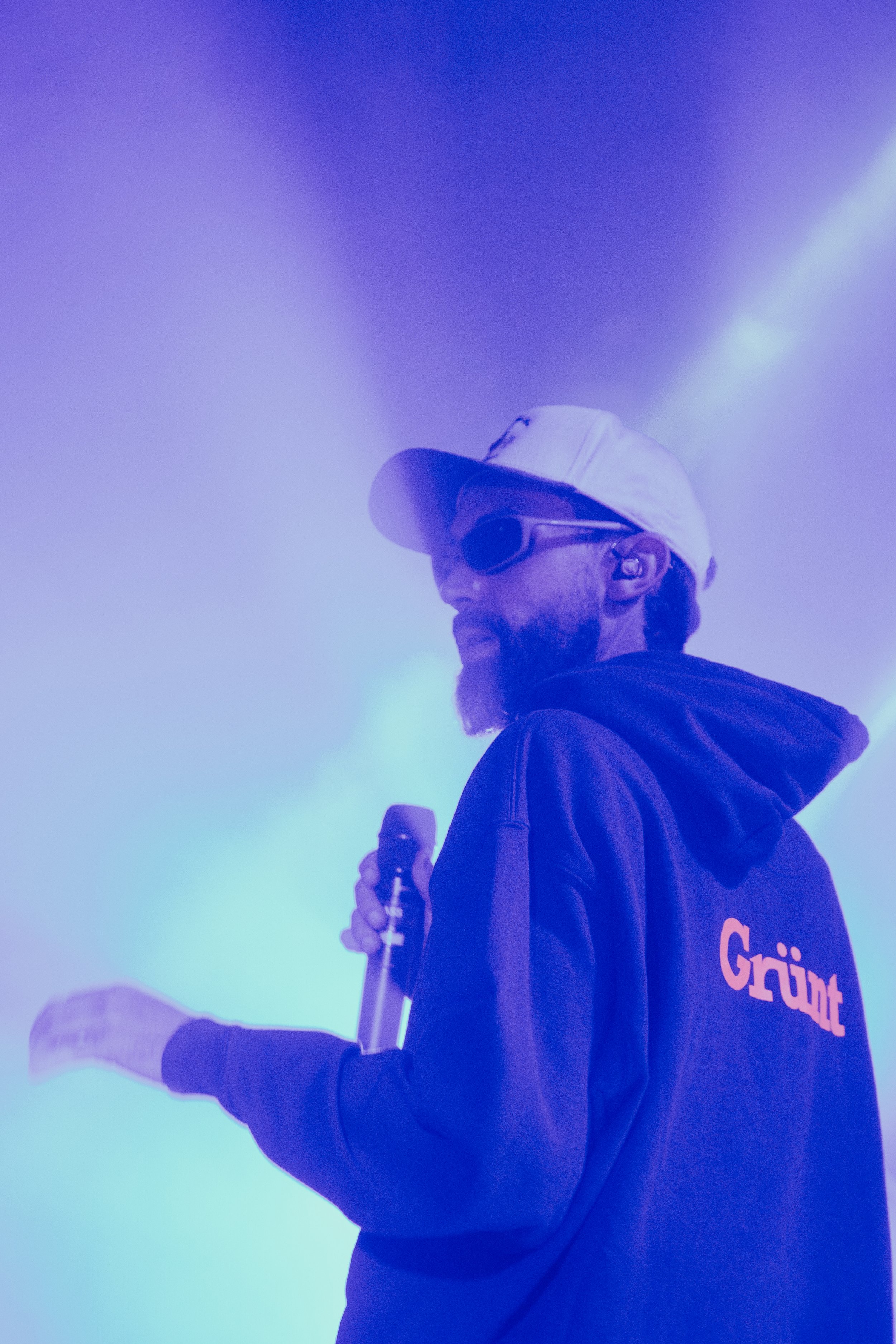 Un homme avec une barbe portant un chapeau blanc, des lunettes de soleil, un sweat à capuche noir avec le mot "Grünt" écrit en orange sur le dos, tenant un microphone dans la main, dans un environnement éclairé par des lumières violettes et bleues.
