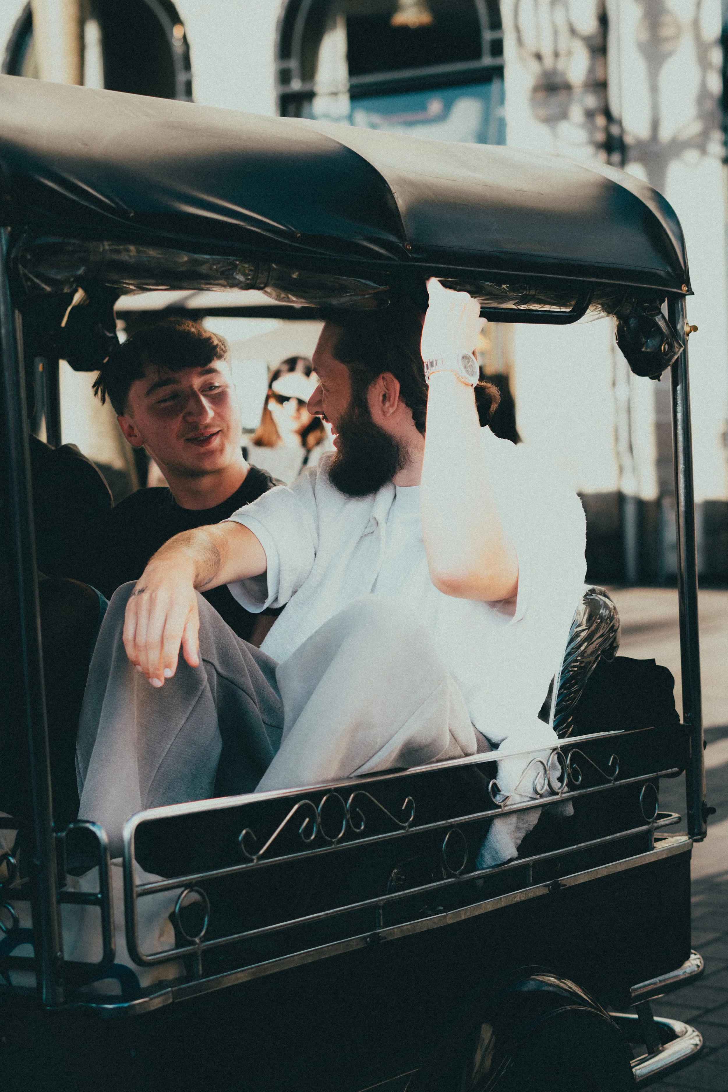 Deux hommes discutent dans une voiturette, l'un a une barbe, l'autre a les cheveux courts, en milieu urbain. La voiturette est noire avec un toit enroulé.