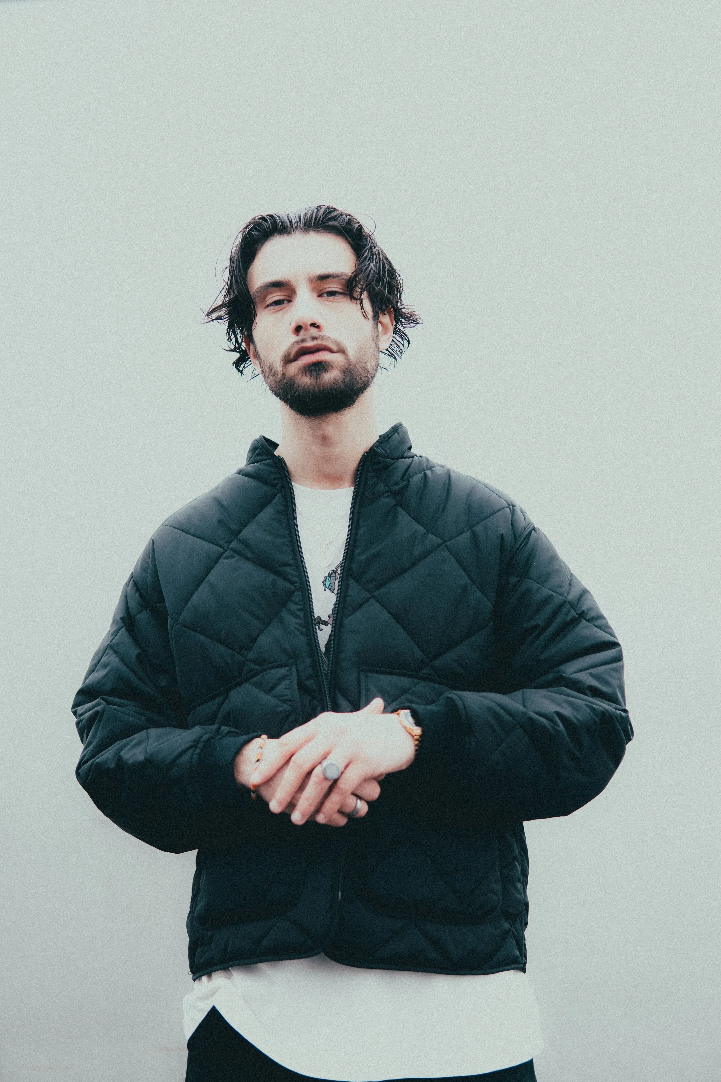 Jeune homme avec des cheveux noirs ondulés, barbe, portant une veste noire puffy, en stance décontractée contre un fond gris uniforme.