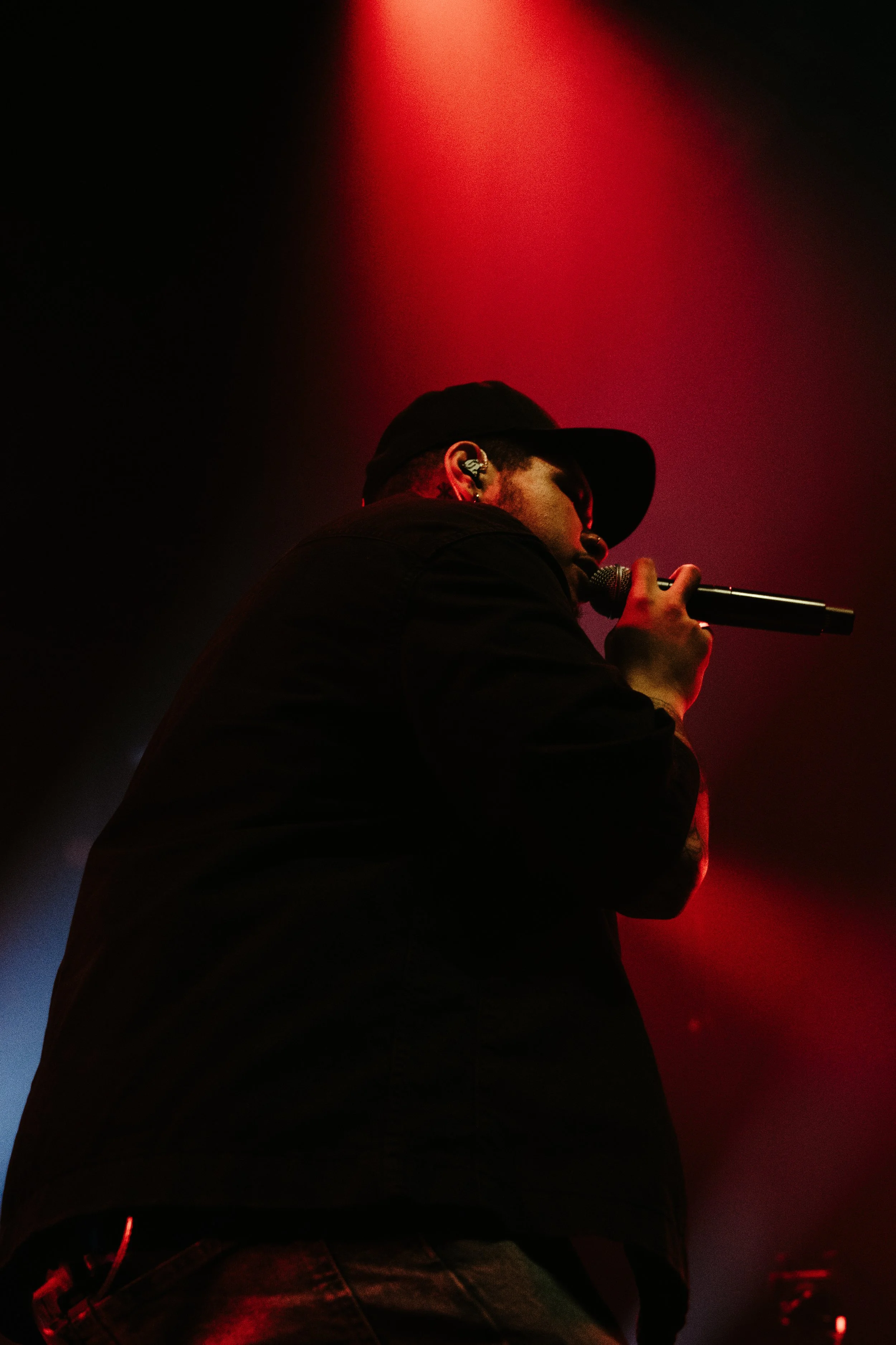 Un chanteur ou rappeur portant une casquette noire, tenant un microphone, illuminé par un éclairage rouge sur scène.