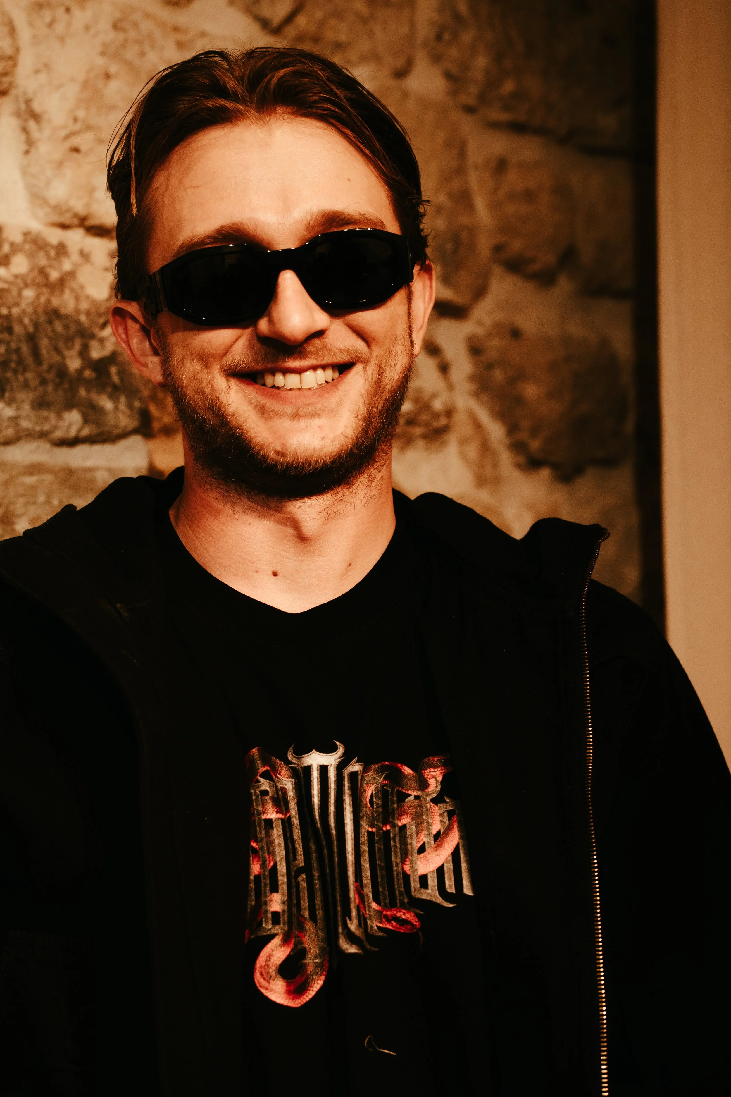 Un homme souriant avec des lunettes de soleil noires, portant un t-shirt noir avec un design de serpent, et un zip noir, posé devant un mur en pierre.