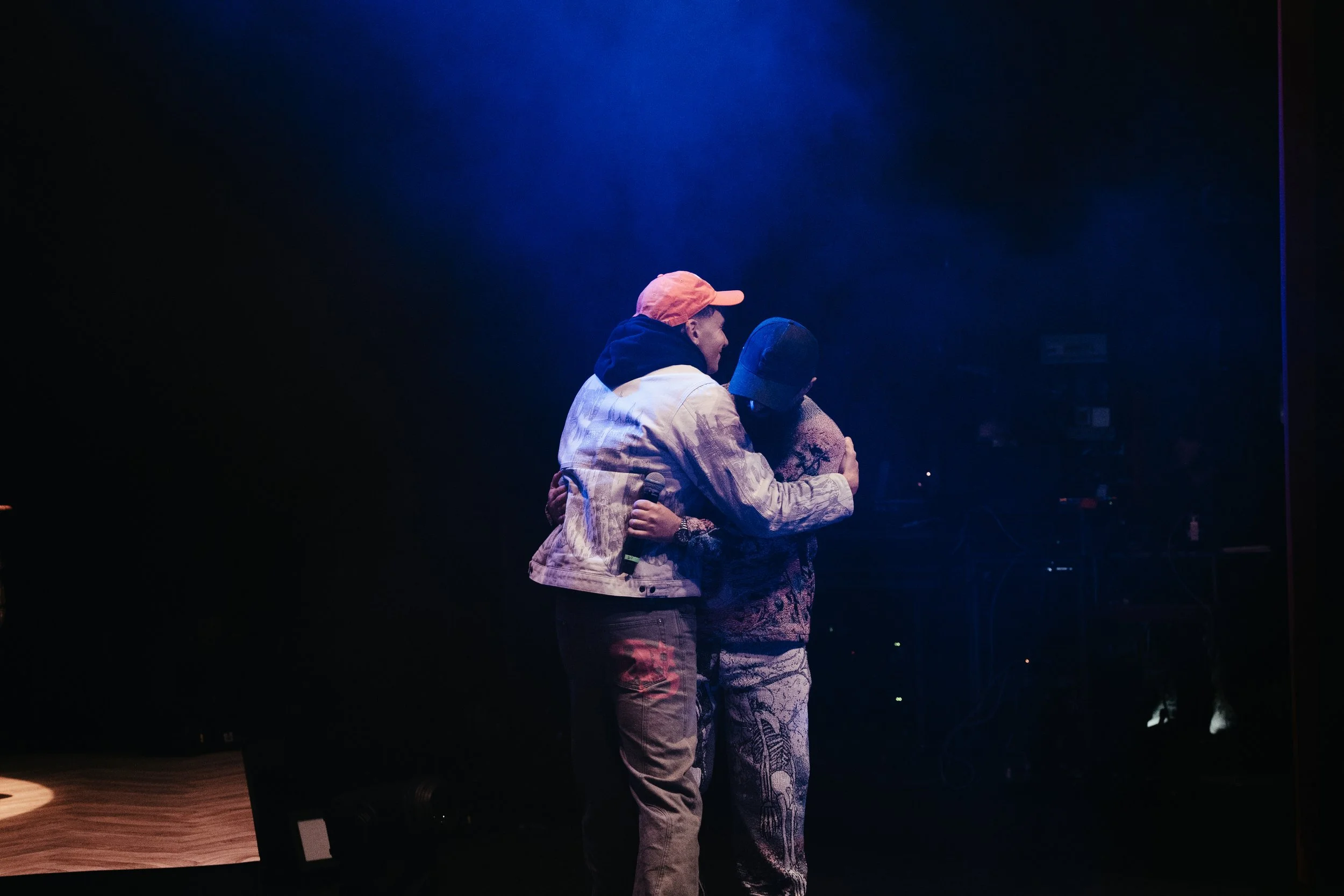 Deux personnes s'embrassent sur scène dans une lumière tamisée, avec le fond sombre et un micro dans une main.