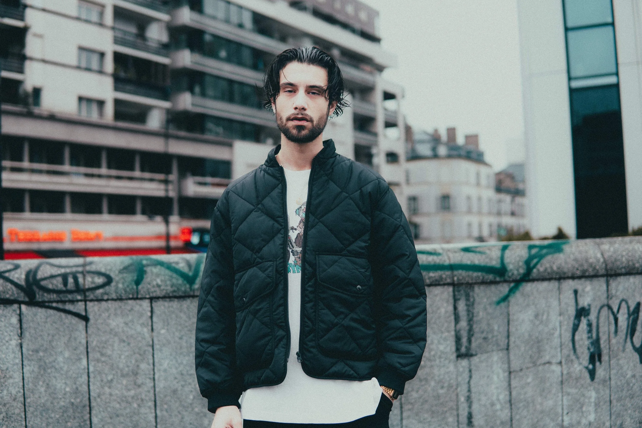 Un homme avec des cheveux noirs et une barbe, portant un manteau noir, se tient devant un mur graffité dans une zone urbaine avec des bâtiments modernes en arrière-plan.