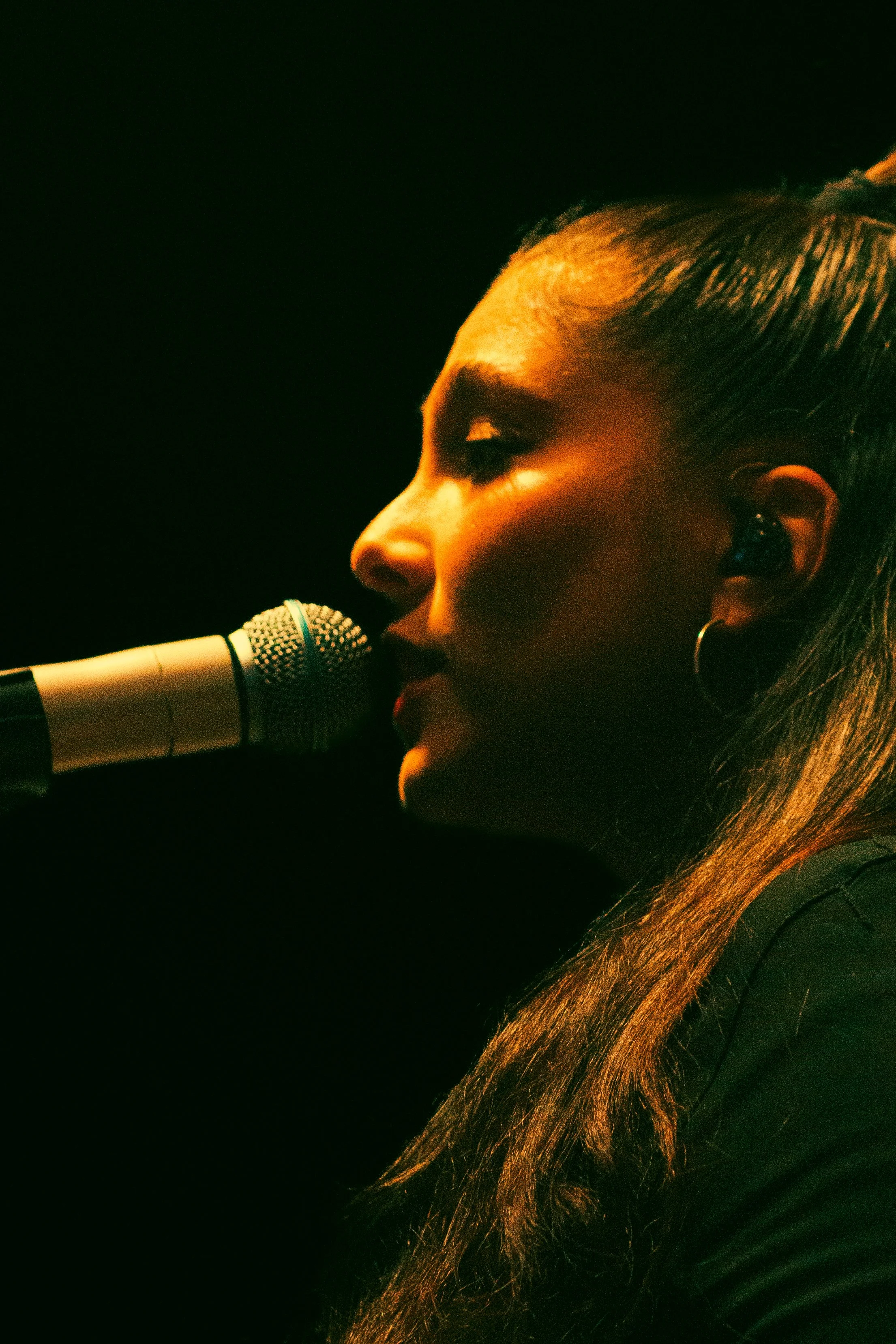 Une femme chante ou parle dans un microphone noir, avec un éclairage chaud et sombre.