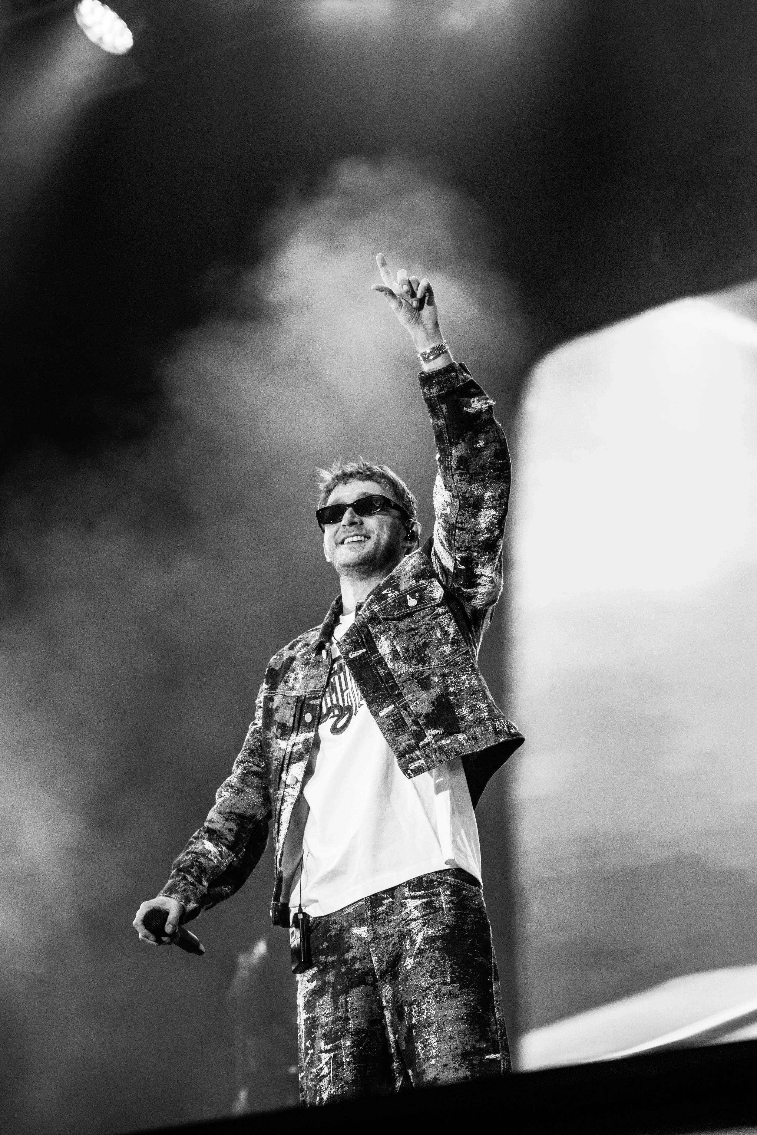 Un homme souriant portant des lunettes de soleil, une veste à rayures et un pantalon à motifs, tient un microphone dans une main et fait un geste avec l'autre main lors d'une performance ou d'un concert, en noir et blanc.