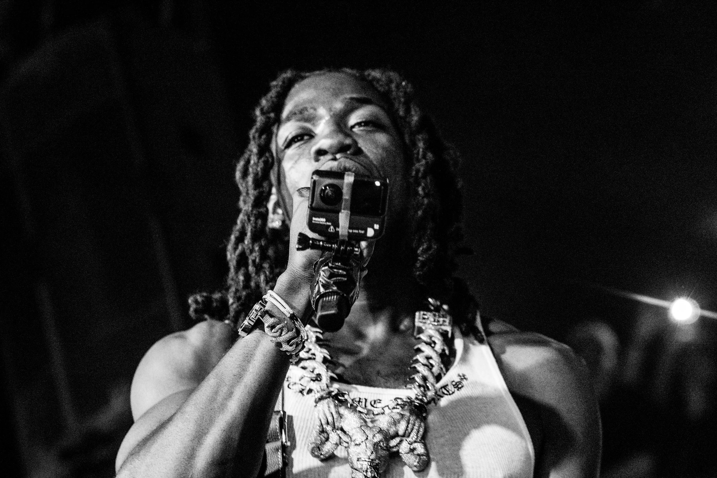 Une femme noire à dreadlocks tient un microphone, portant une chaîne avec une décoration en forme de lion, un tank top blanc et des bracelets, prise dans un style noir et blanc.
