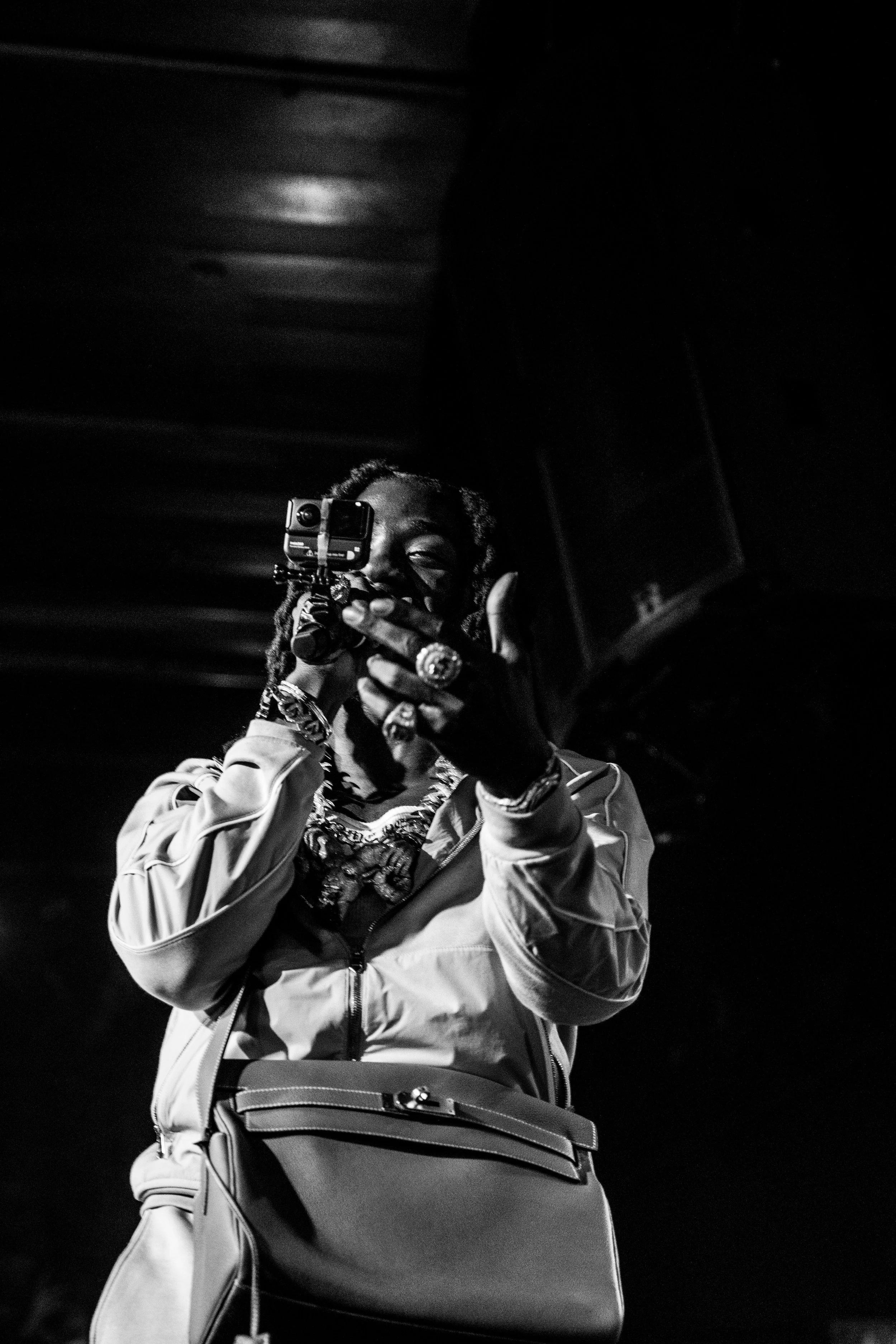 Une personne portant des bijoux, habillée d'une veste avec une sacoche, tient un appareil photo ou une caméra et semble en train de prendre une photo ou de filmer.