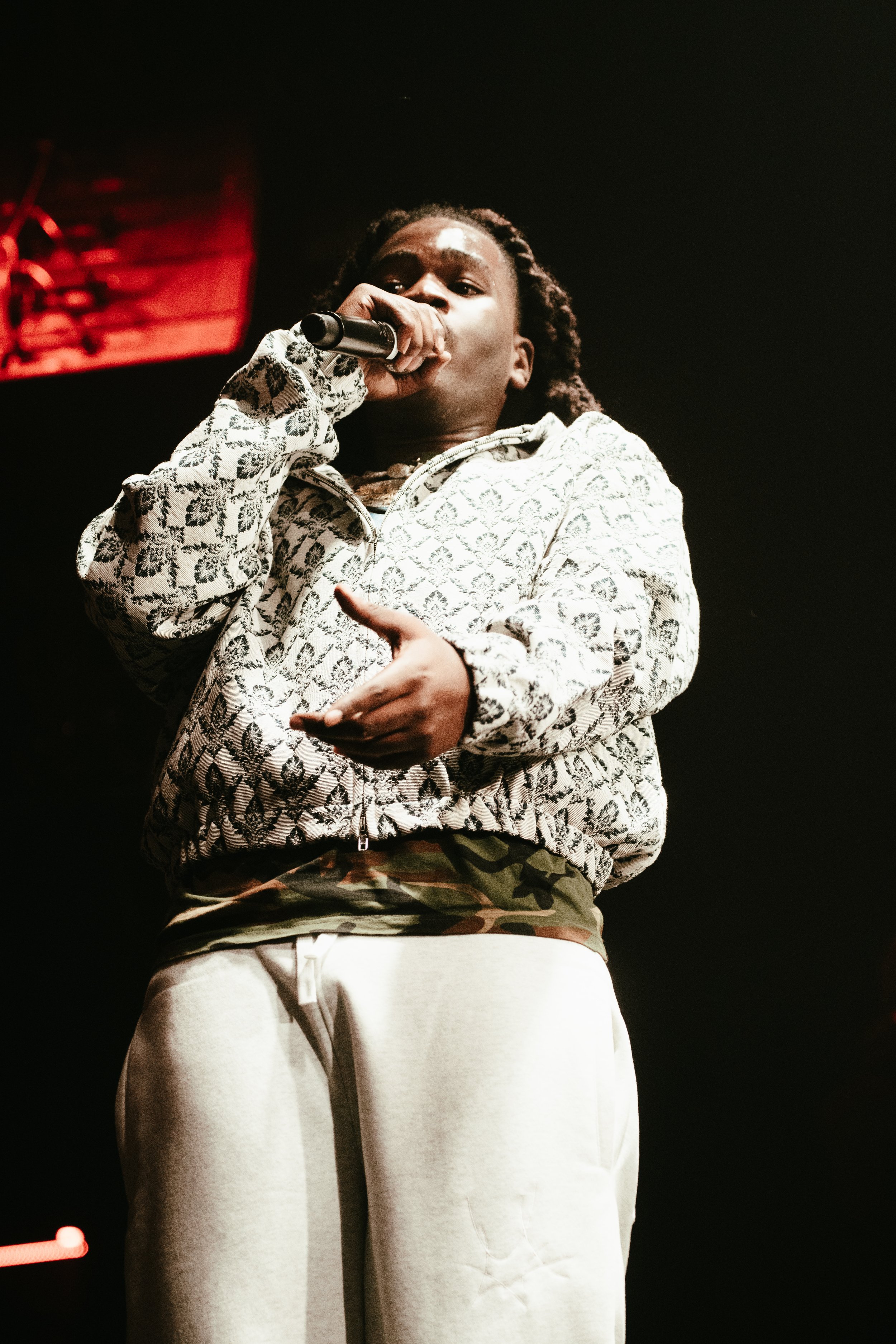 Une personne tenant un micro en train de faire un performance, vêtue d'une veste à motif blanc et noir, avec un fond sombre.