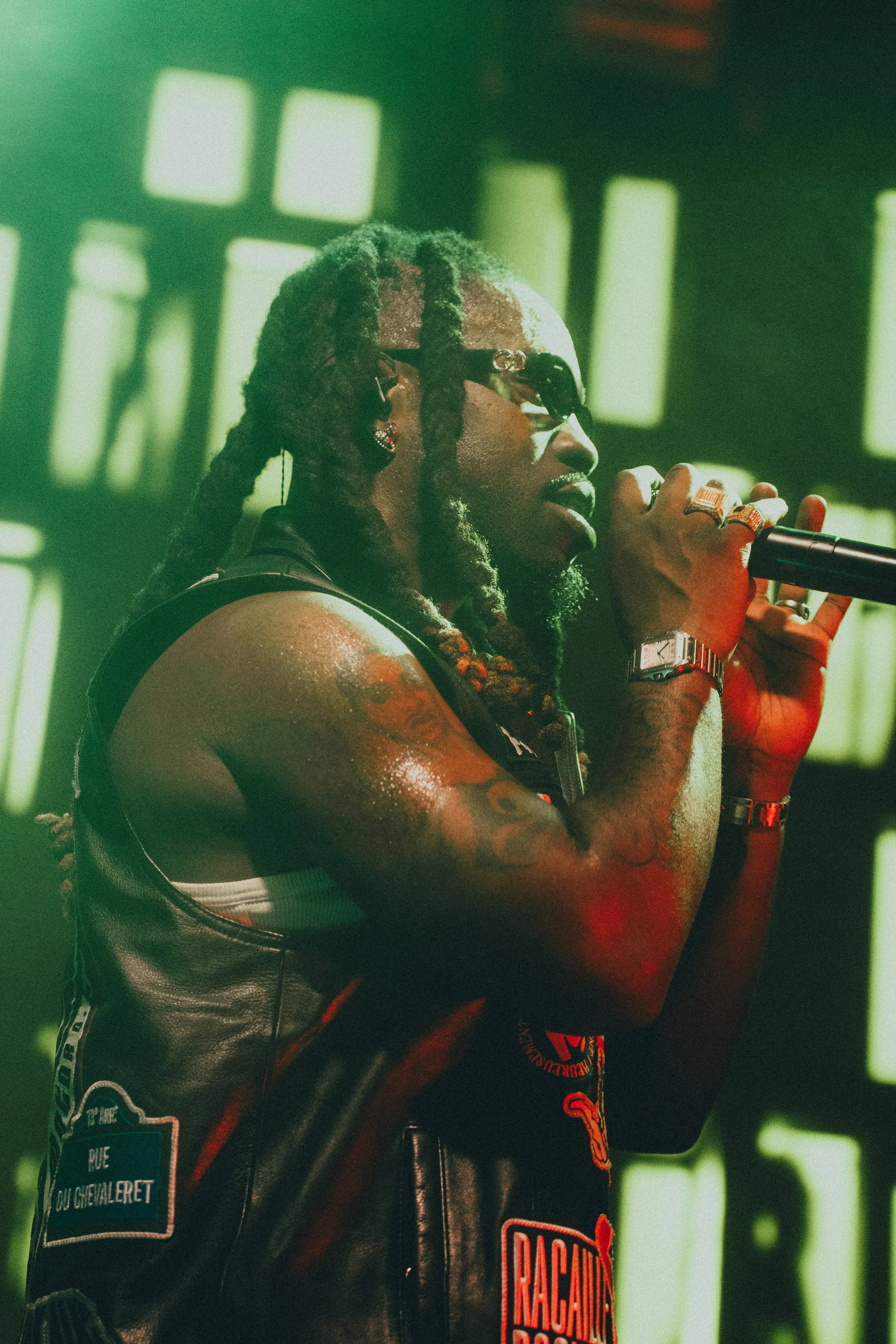 Un homme avec des dreads, portant des lunettes de soleil, chantant dans un microphone sur scène, portant une veste en cuir avec des patches, sous un éclairage vert.