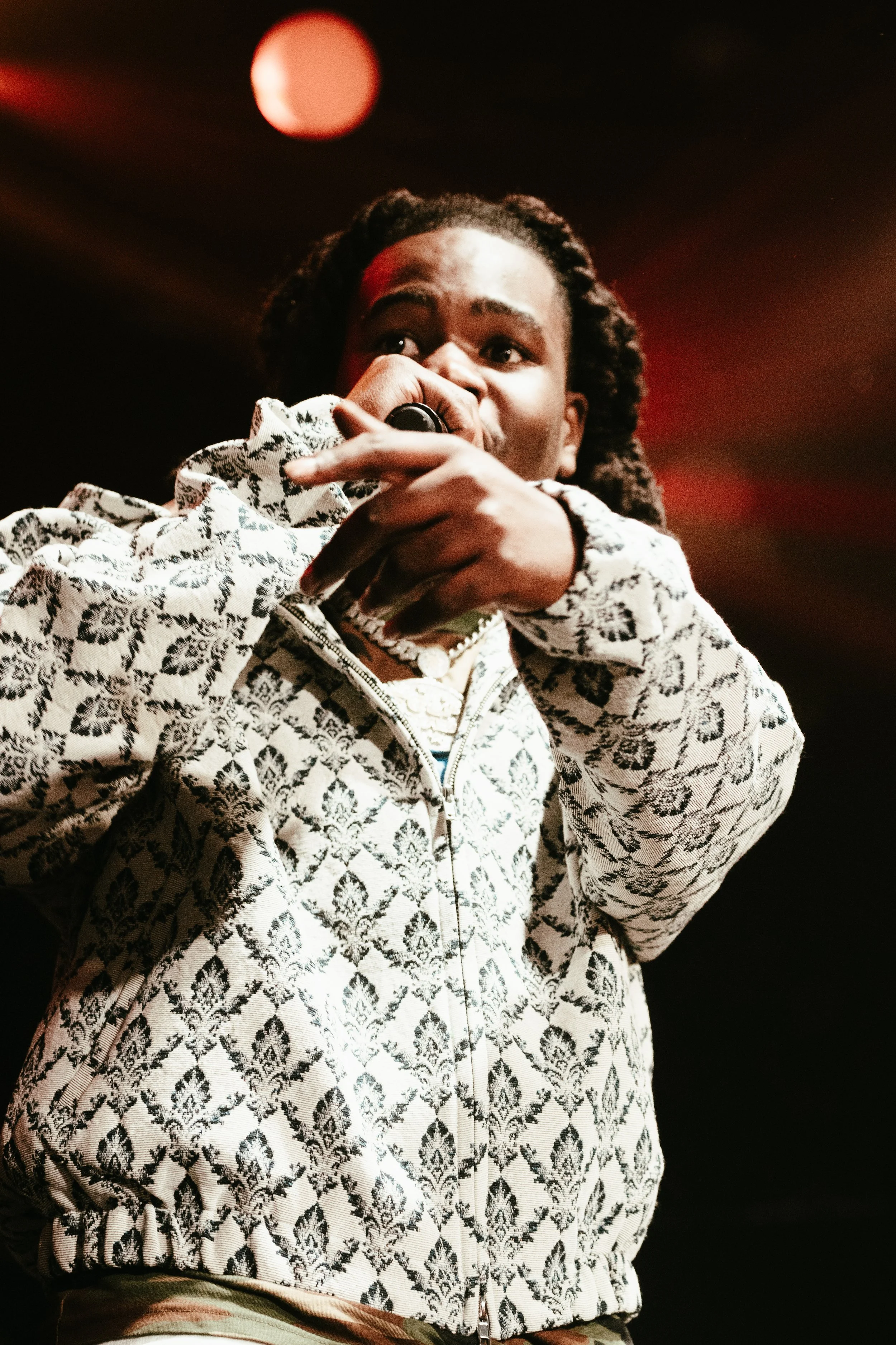 Un artiste de rap en pleine performance, tenant un micro, portant une veste imprimée à motifs noirs et blancs, sous un éclairage chaud, avec un fond sombre et une lumière orange en arrière-plan.