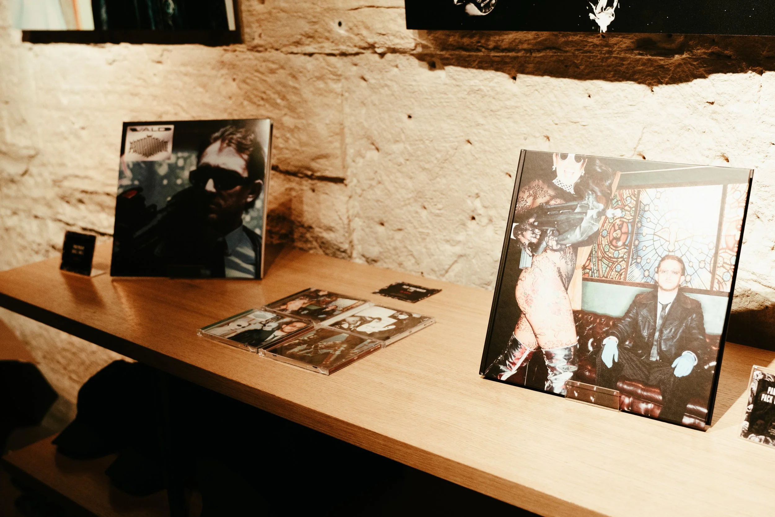 Exposition de plusieurs albums et photos sur une table en bois, contre un mur en pierre beige. Deux albums avec des images de personnes, dont une avec une femme en vêtement noir et un homme assis en costume, dans une salle avec vitraux.