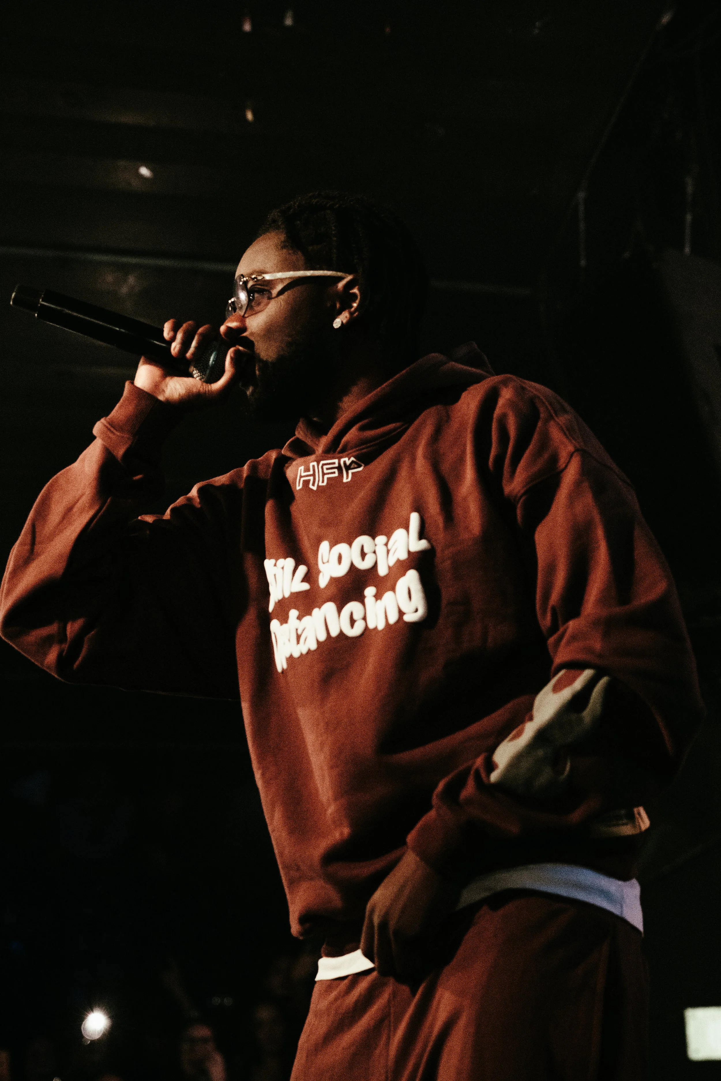 Un homme avec des dreadlocks portant des lunettes, une boucle d'oreille, et un sweat à capuche descend de l'épaule avec un texte qui dit 'HIZ SOCIAL Distancing'. Il tient un microphone à la main dans un environnement sombre, apparenté à une scène ou 