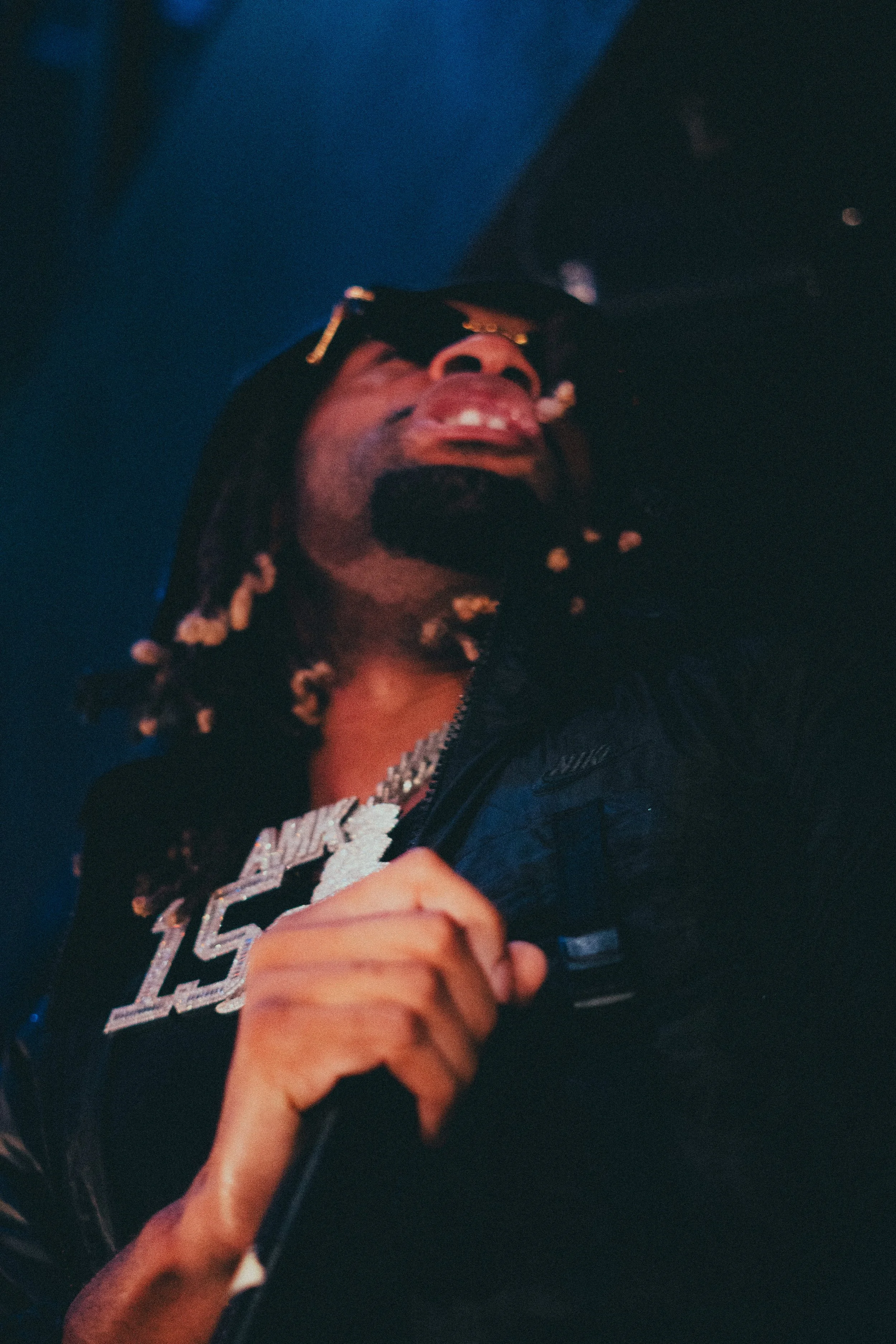 Un homme portant des lunettes de soleil et chaîne en diamant tenant un microphone lors d'une performance ou d'un concert, sous un éclairage sombre.