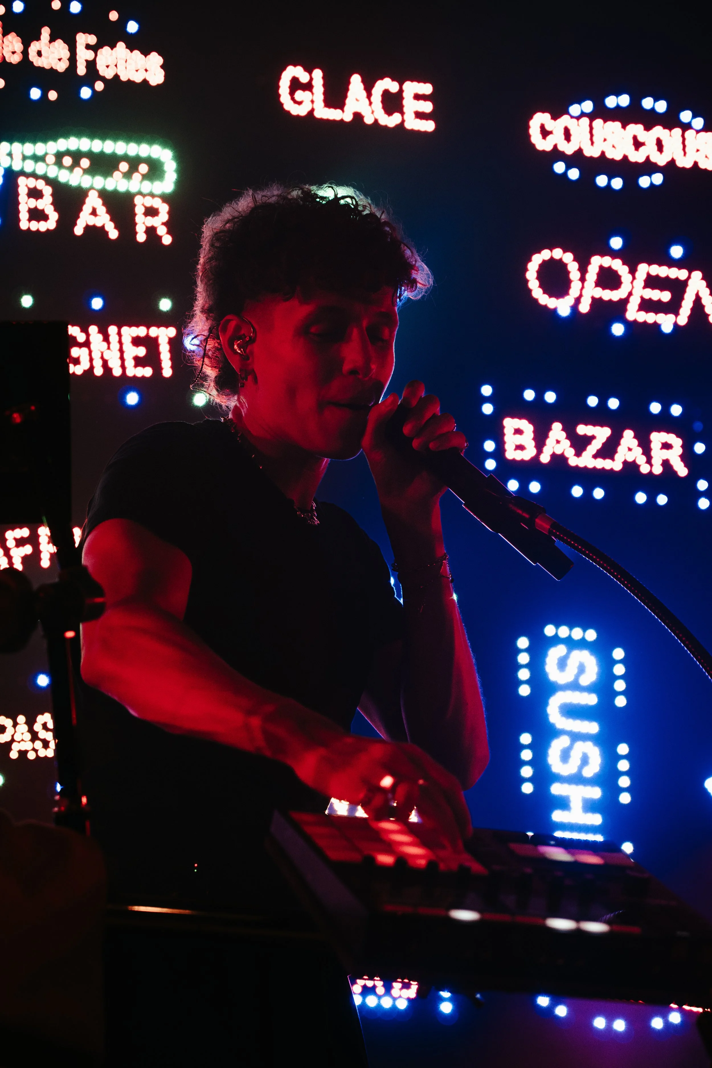 Une femme chante et joue avec un clavier face à un fond de néons lumineux avec des mots en français et en anglais tels que 'GLACE', 'COULEURS', 'OPEN', 'BAZAR', 'SUSHI'.