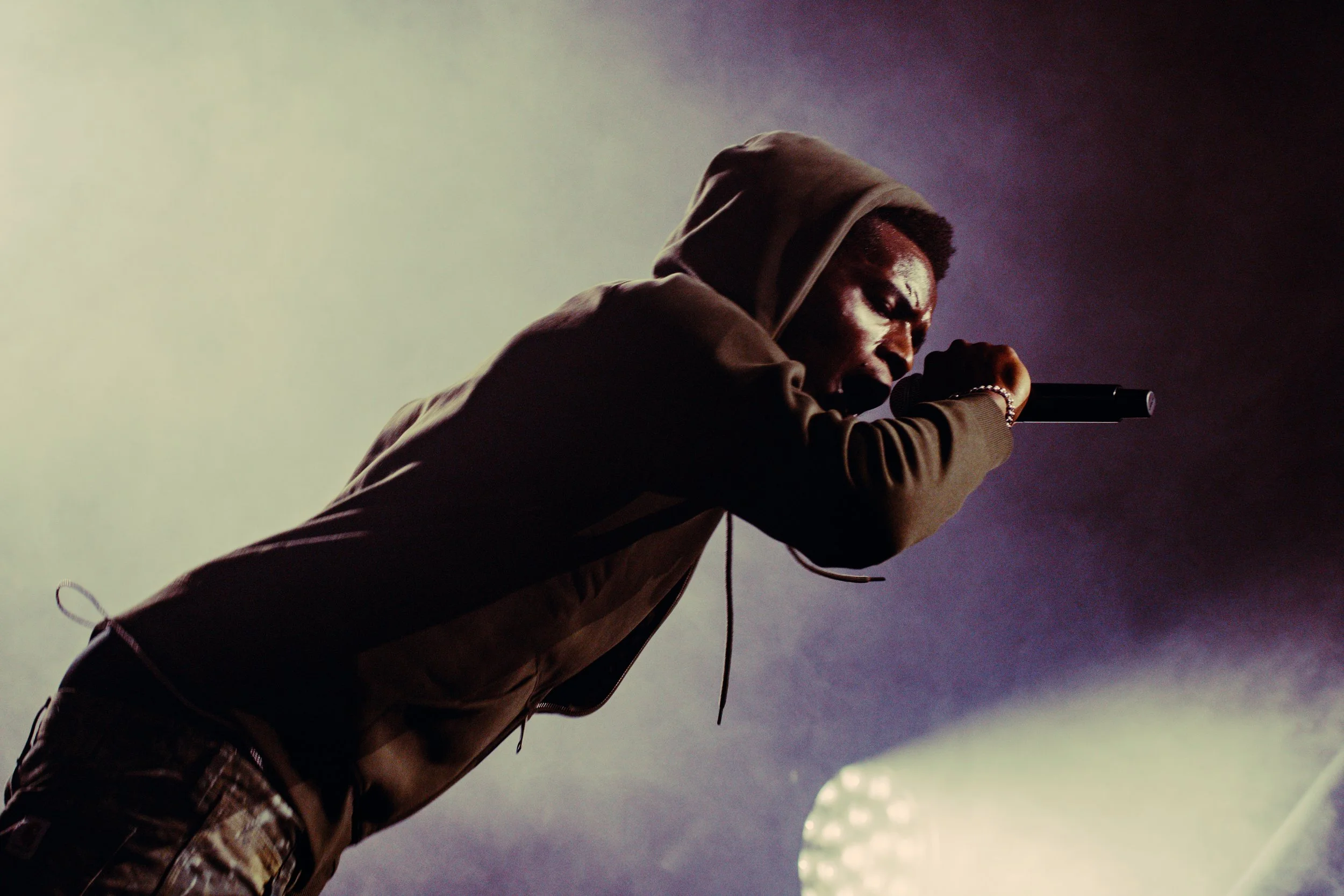 Un homme tenant un microphone prêt à chanter ou à parler, vêtu d'une veste à capuche, dans un environnement sombre et brumeux.