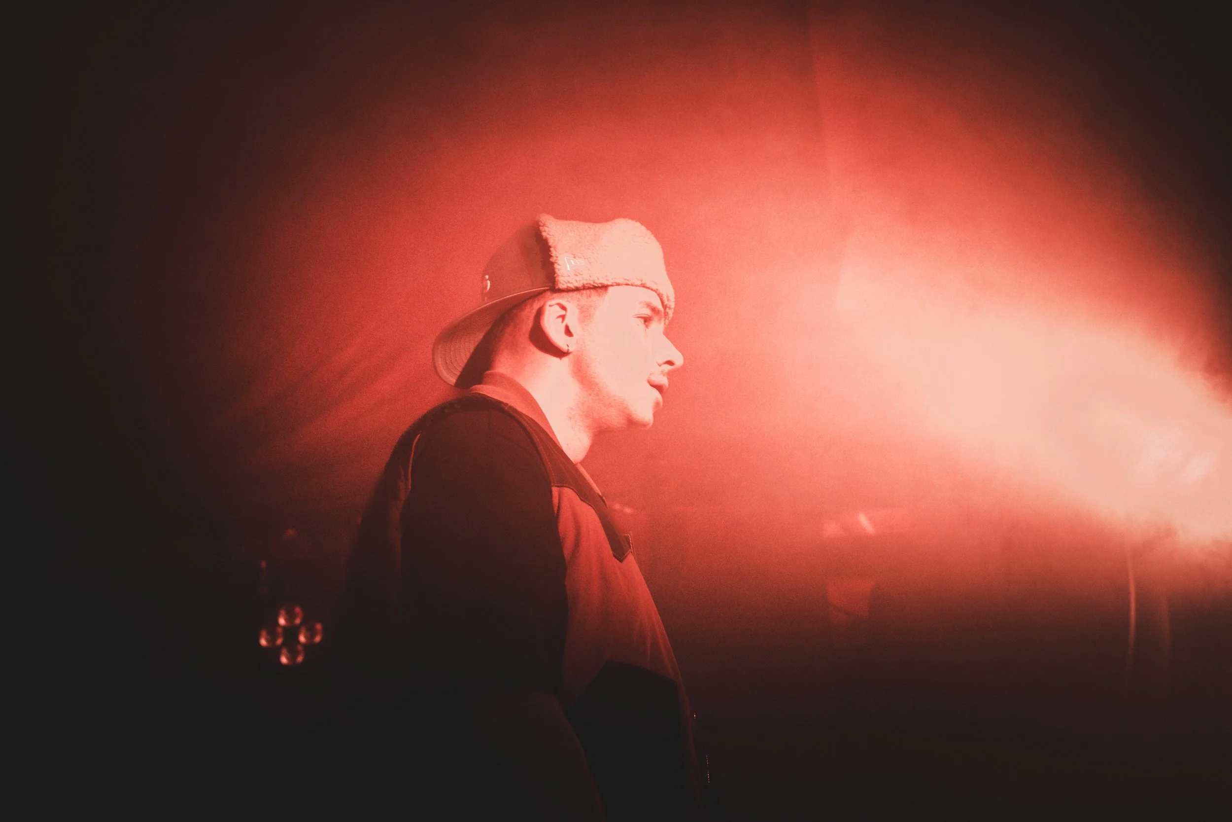 Un jeune homme porte un bonnet blanc et une veste noire, entouré de lumière rose/orange dans une ambiance sombre.