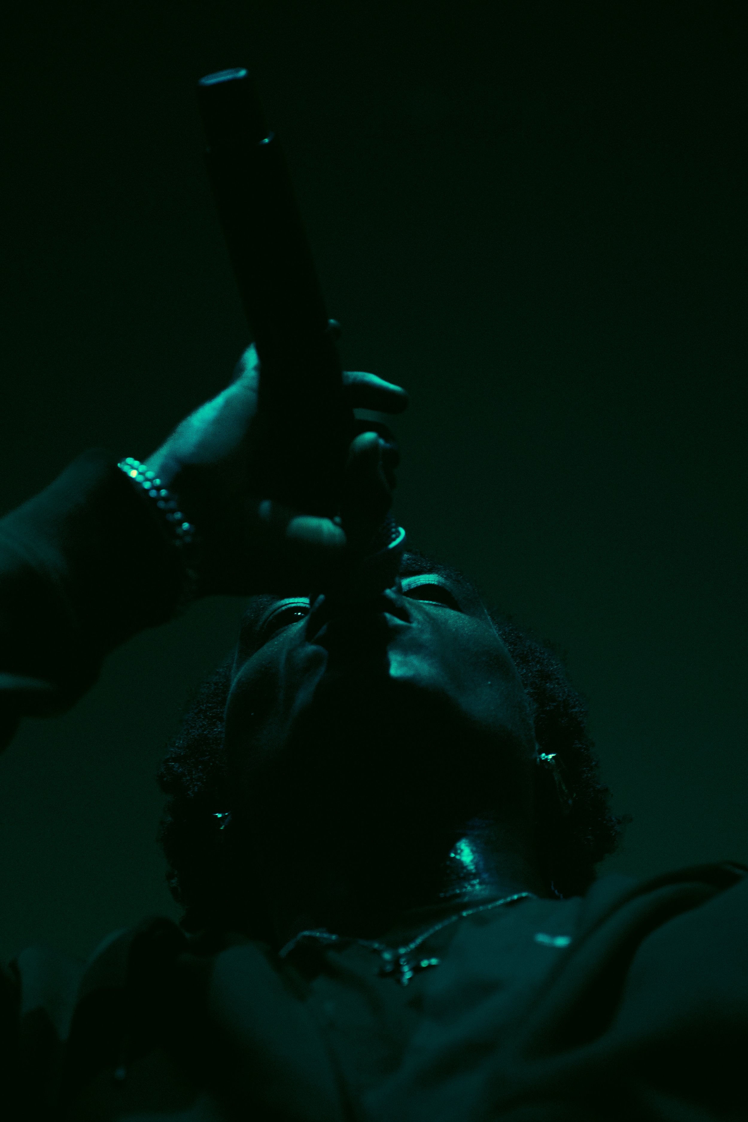 Une personne noire portant des bijoux et une veste noir, vue de dessous, tenant un pistolet pointé vers le haut dans un éclairage sombre.