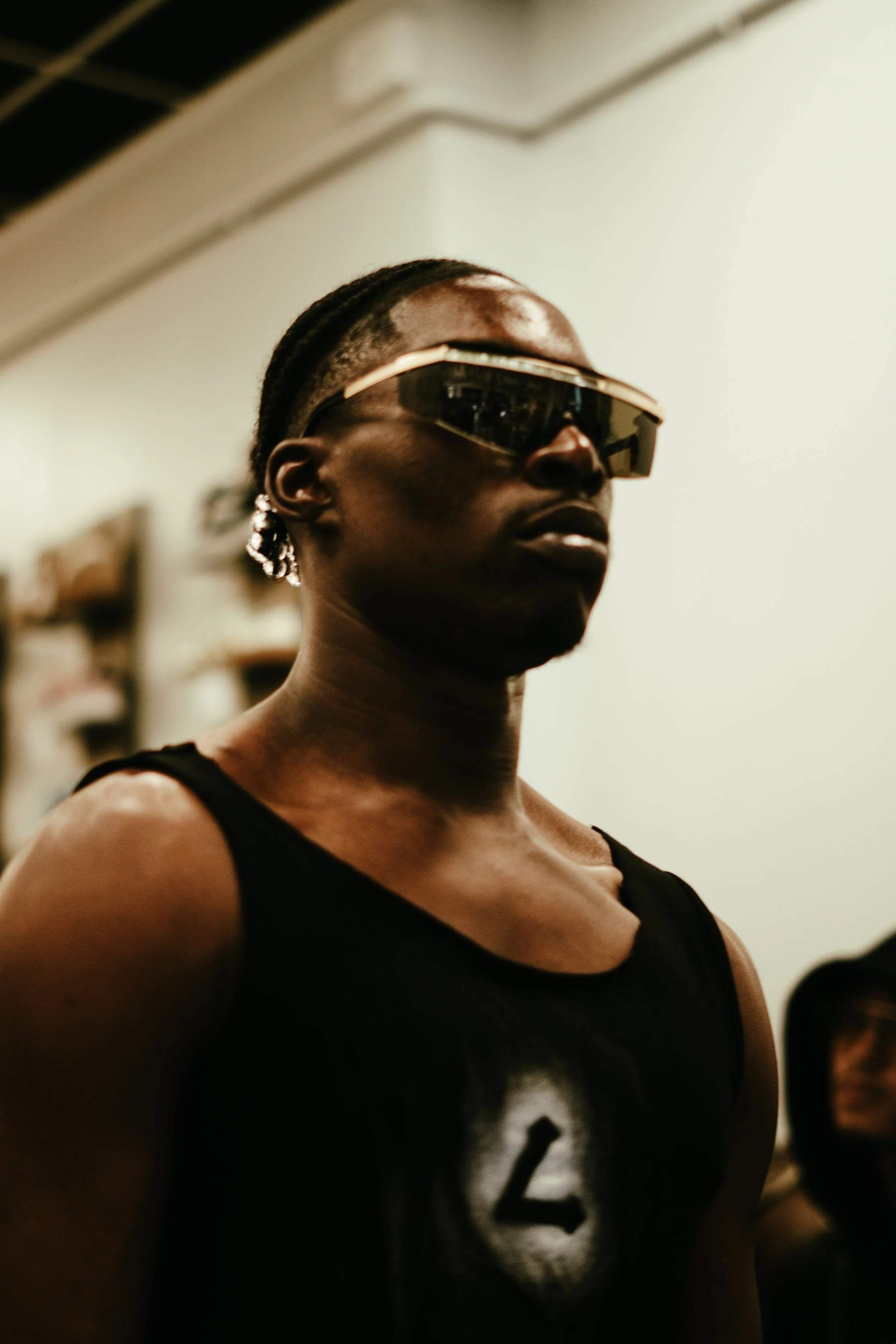 Jeune femme portant des lunettes de soleil futuristes, un haut noir sans manches avec un logo et des boucles d'oreilles en forme de crocodile, dans un environnement intérieur.