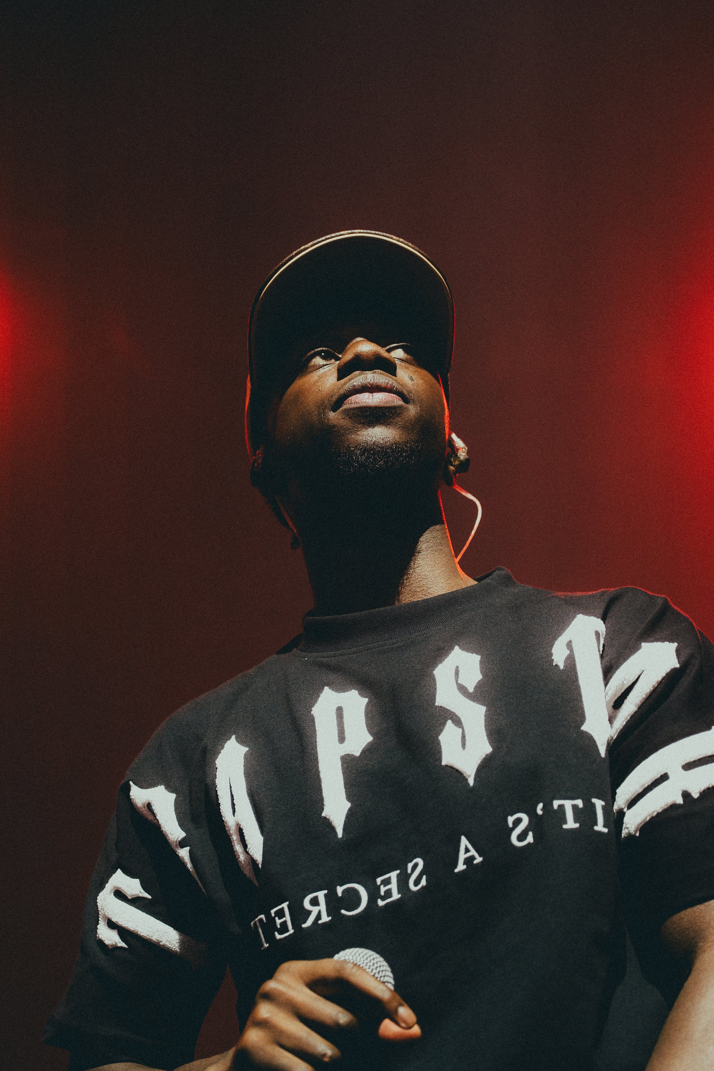 Un homme portant une casquette et un t-shirt noir avec des inscriptions blanches, tenant un microphone, semble être en train de performer sur scène sous un éclairage rouge.