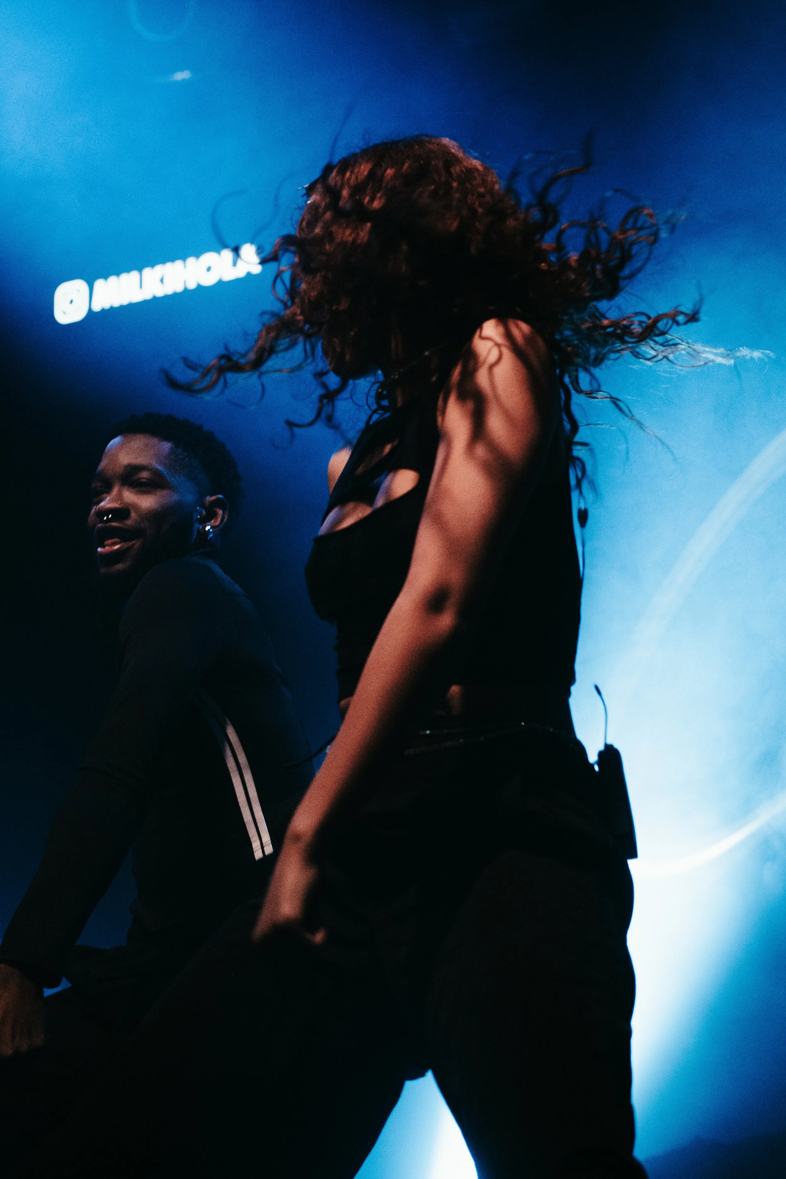 Deux personnes dansant sous des lumières bleues, une aux cheveux bouclés, l'autre avec des cheveux courts, lors d'une performance ou d'un concert.