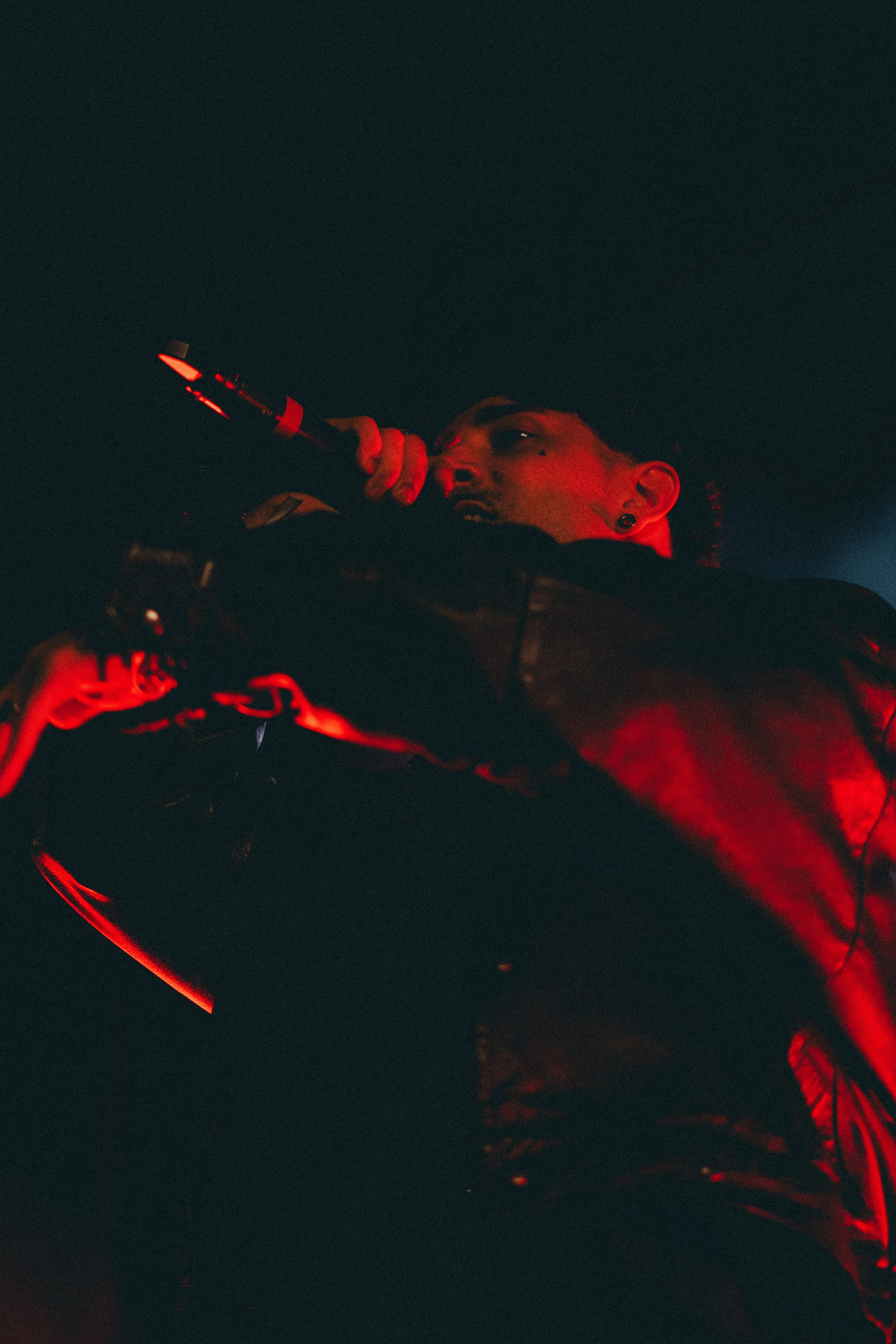 Un homme qui chante ou parle dans un microphone, éclairé en rouge, portant une veste en cuir, dans un environnement sombre ou une scène.