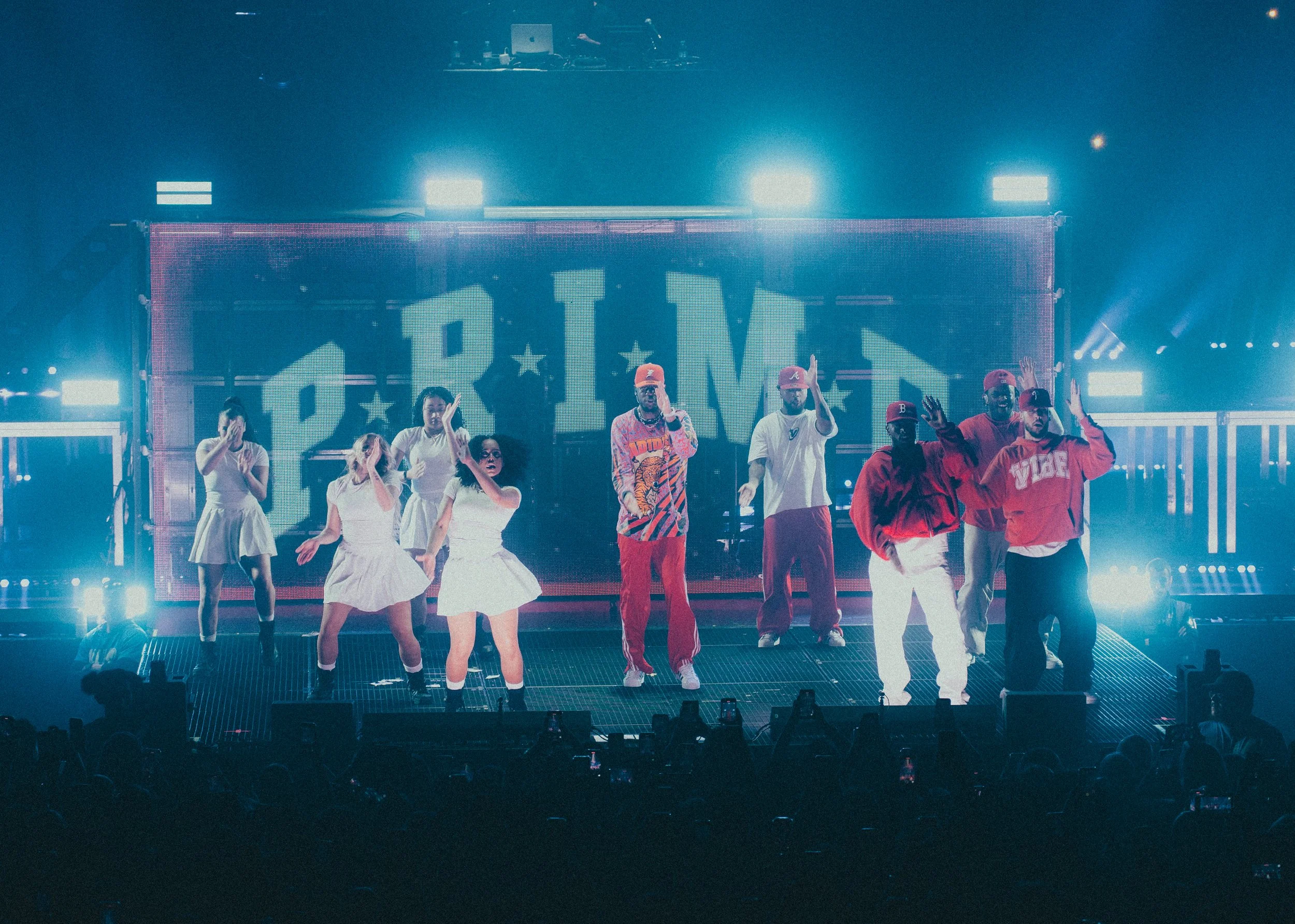 Groupe de personnes sur scène lors d'un concert, avec un grand écran en arrière-plan affichant le nom 'PRIME'.
