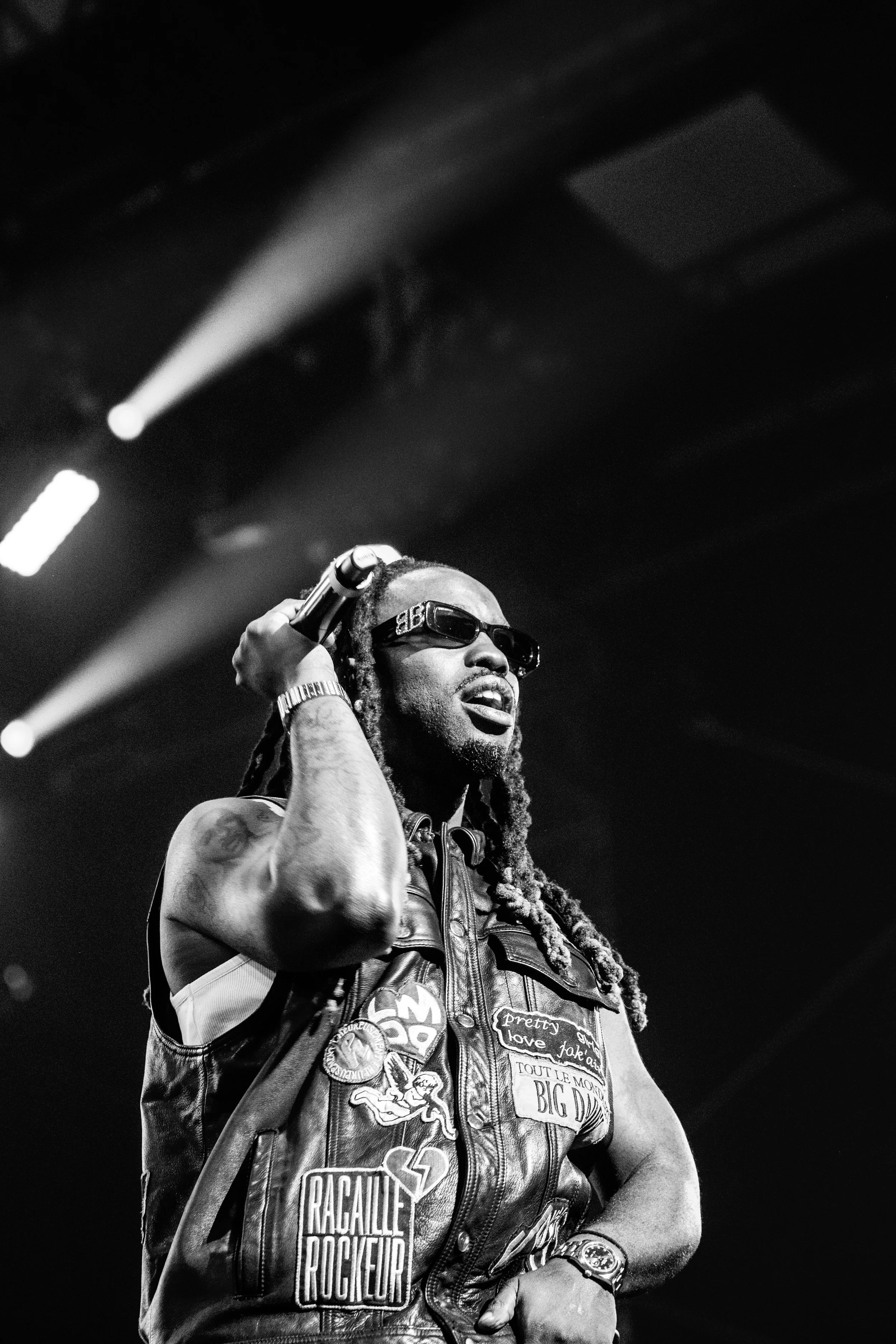 Un homme avec des dreadlocks, portant des lunettes de soleil et une veste en cuir, chante ou parle lors d'un concert, sous des lumières de scène.