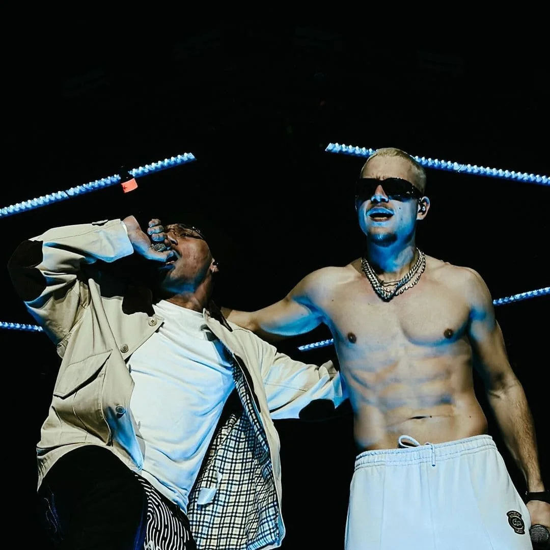Deux hommes, l'un chantant avec un micro, l'autre en maillot de bain, portant des lunettes de soleil, sous un éclairage bleu.