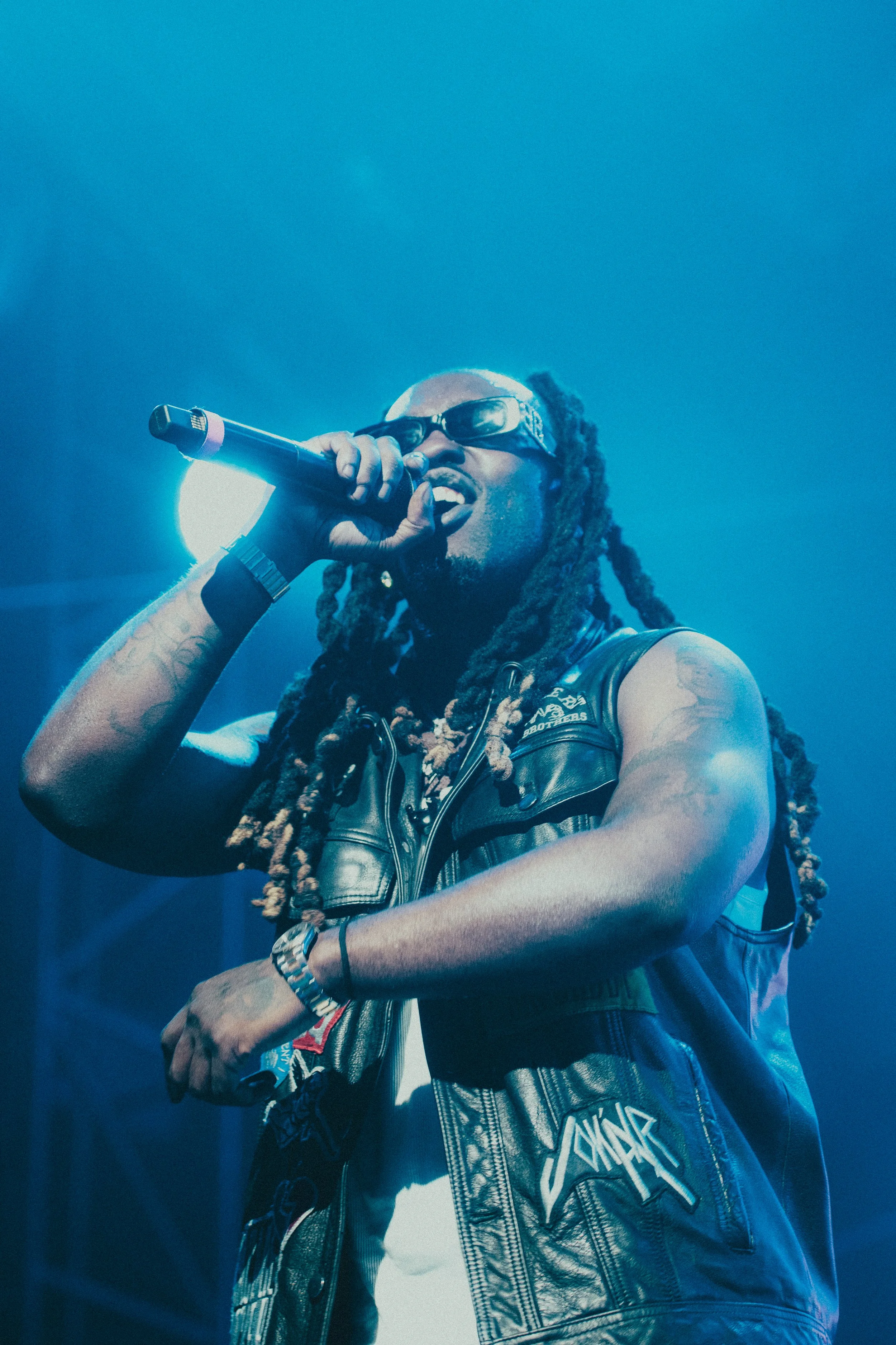 Un homme portant des lunettes de soleil, une veste en cuir, et tenant un microphone en pleine performance sur scène