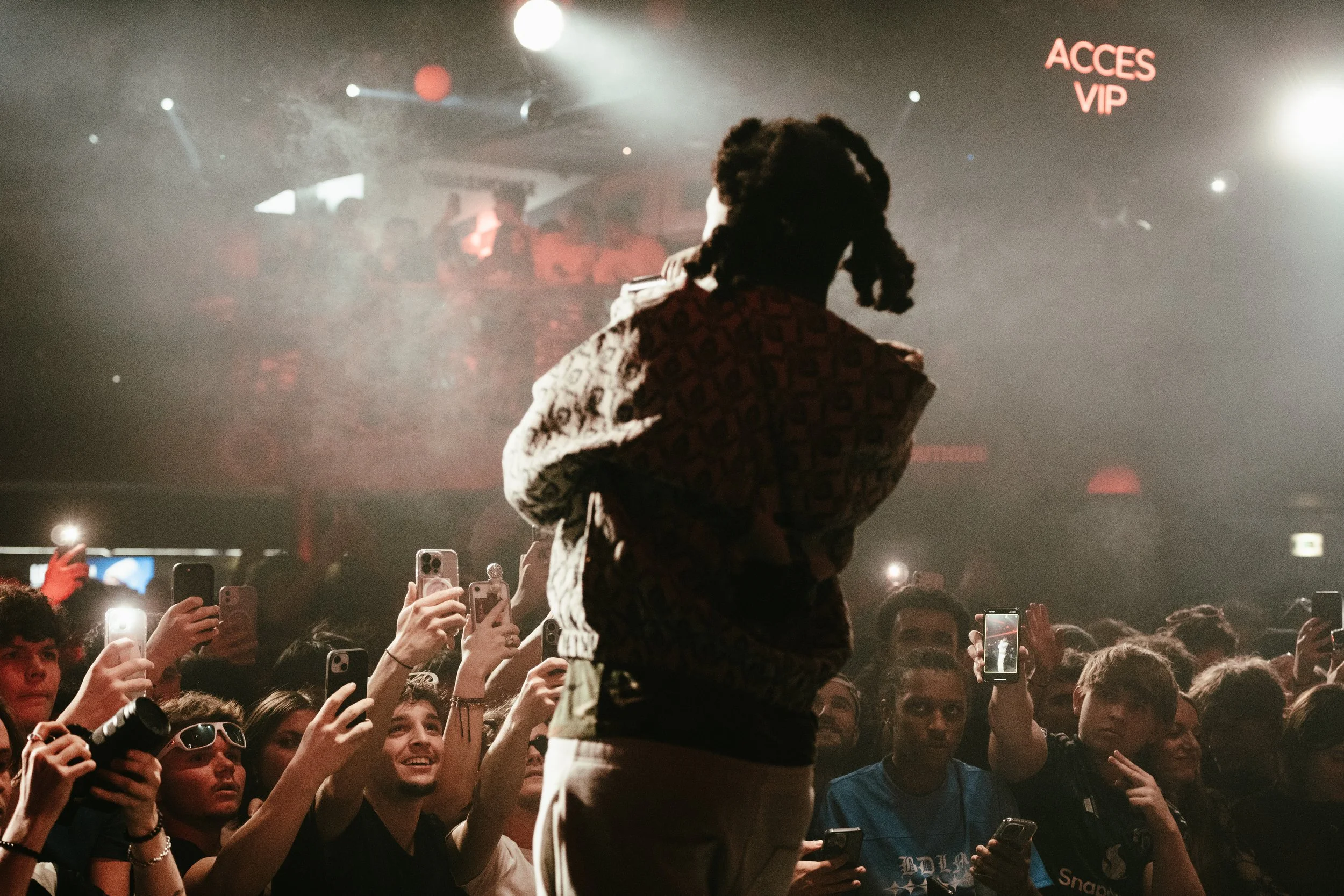 Un artiste avec des dreadlocks rencontre un public nombreux dans une salle de concert, certains spectateurs prennent des selfies ou des vidéos avec leurs téléphones.