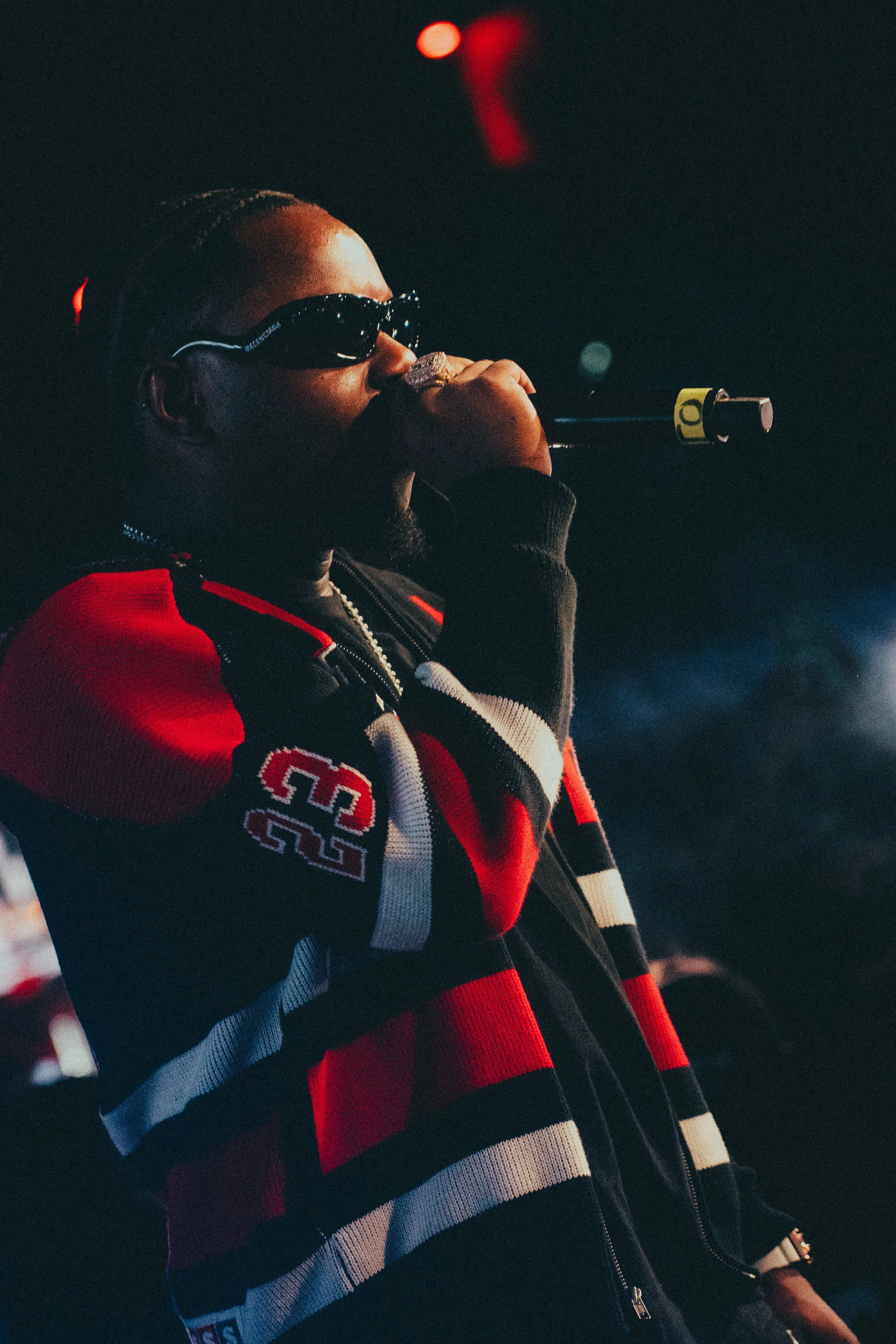 Un artiste portant des lunettes de soleil noires, avec une barbe, qui chante ou parle dans un microphone lors d'une performance, vêtu d'un sweat à rayures rouges, noires, blanches, avec le numéro 23.