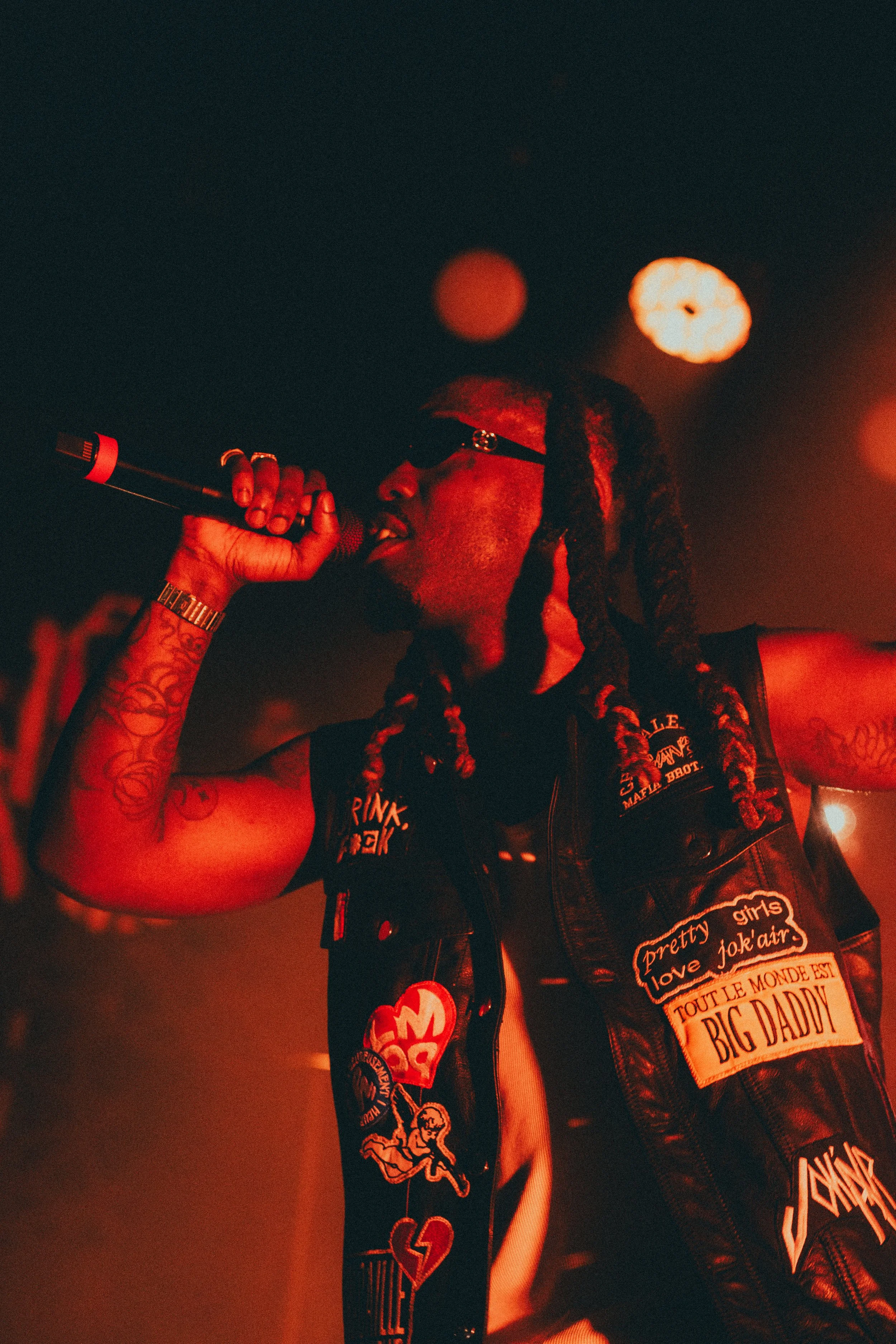 Un homme noir avec des dreadlocks, portant des lunettes de soleil et un gilet en cuir avec plusieurs patchs, chante dans un micro lors d'une performance musicale, sous un éclairage rouge.