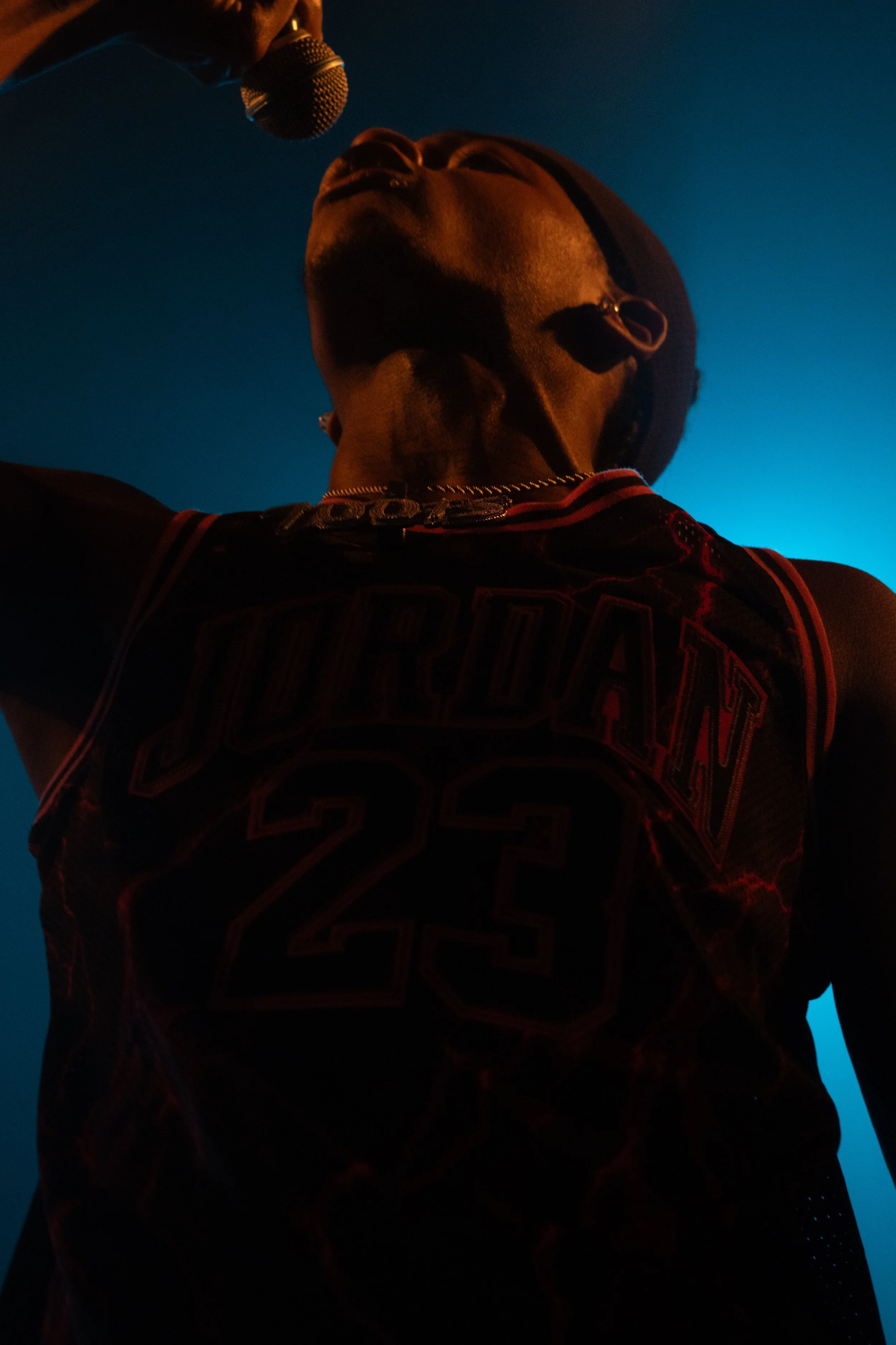 Une personne portant un maillot de basketball avec le nom 'JORDAN' et le numéro 23, vue de dessous avec un micro suspendu au-dessus, fond bleu foncé.