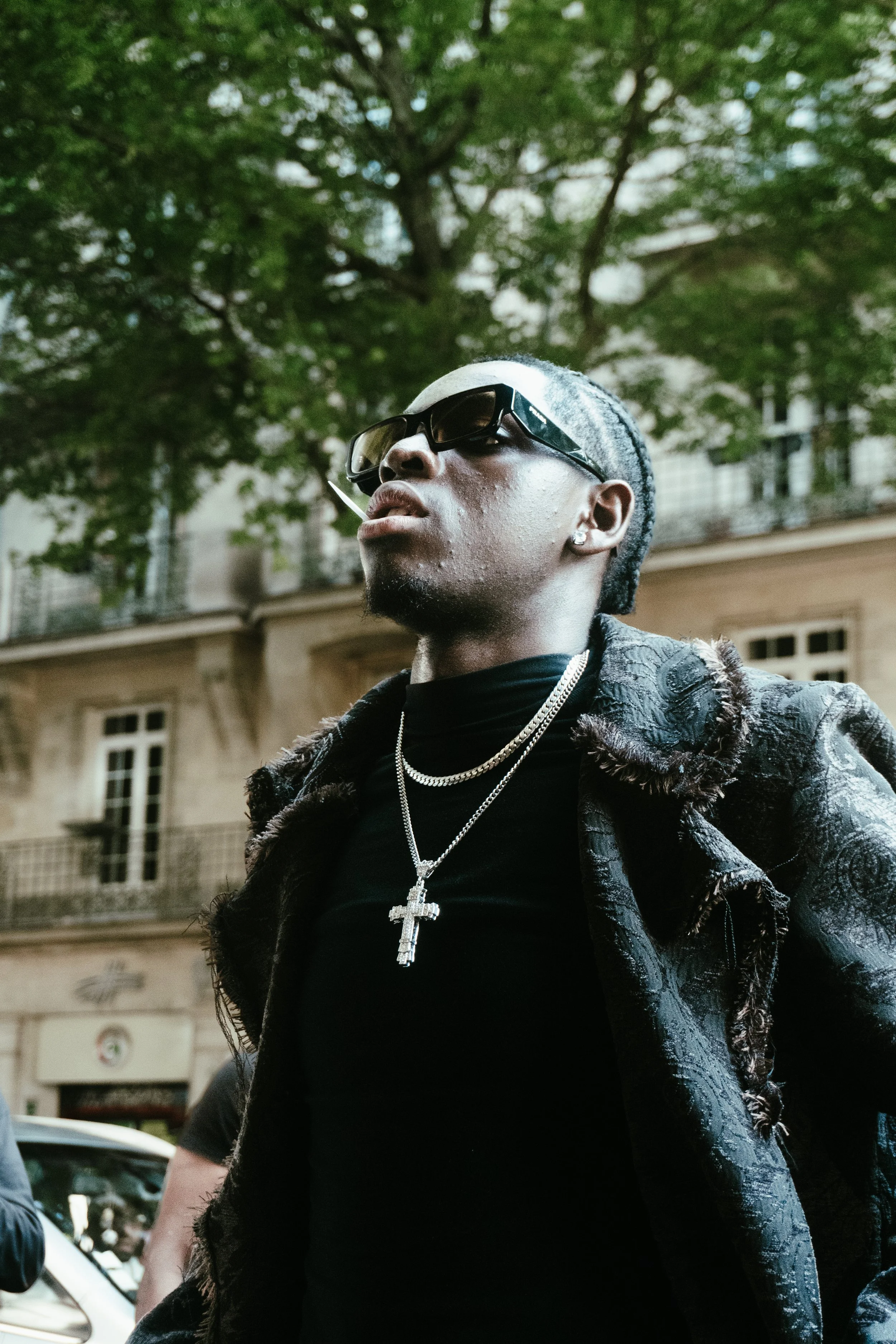Un homme portant des lunettes de soleil, un collier avec une croix, une veste en tissu foncé et une tenue noire, se tient devant un bâtiment et un arbre feuillu.