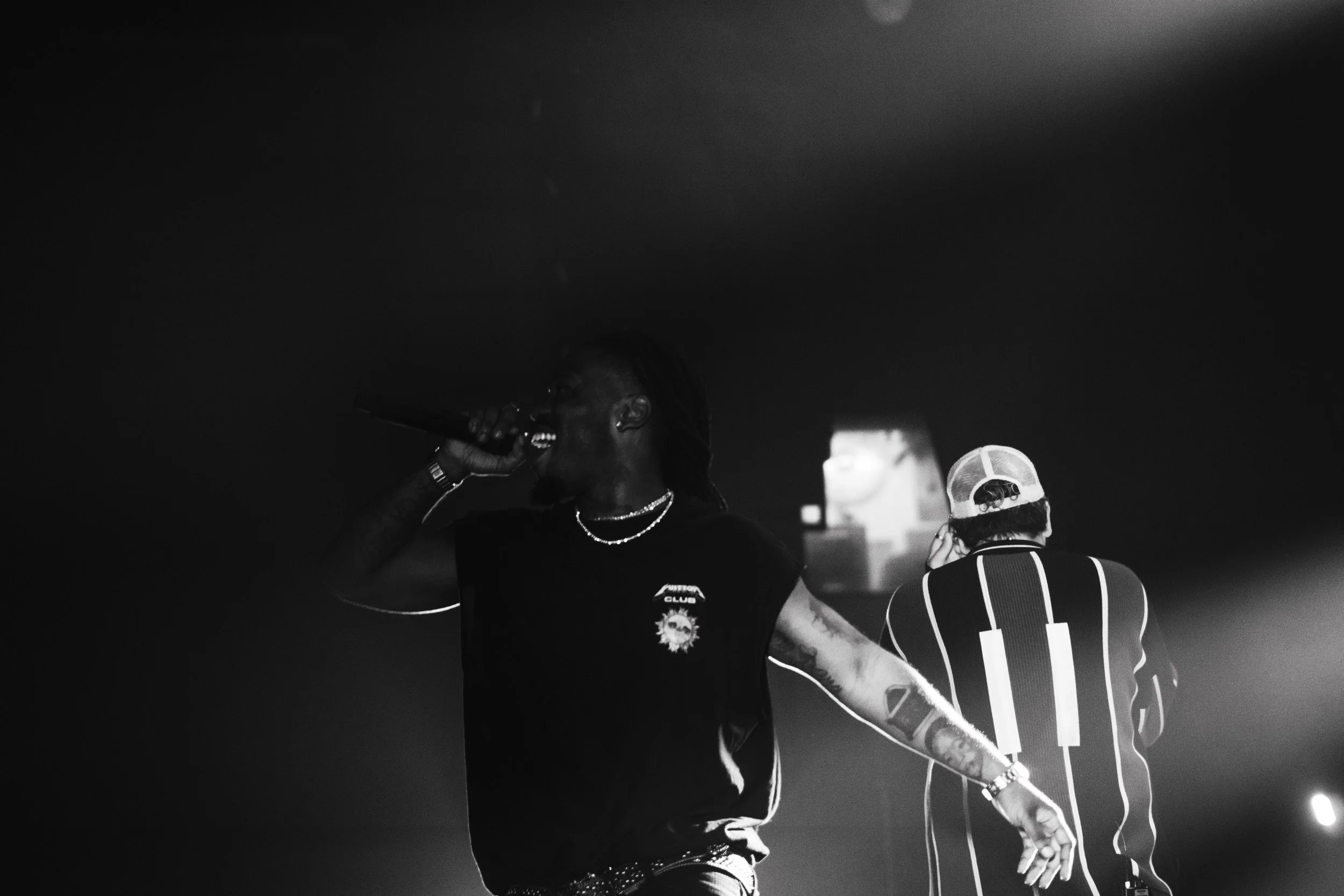 Deux chanteurs sur scène, un chanteur avec un micro, en t-shirt noir et un autre avec un chapeau et des vêtements rayés, dans une ambiance de concert