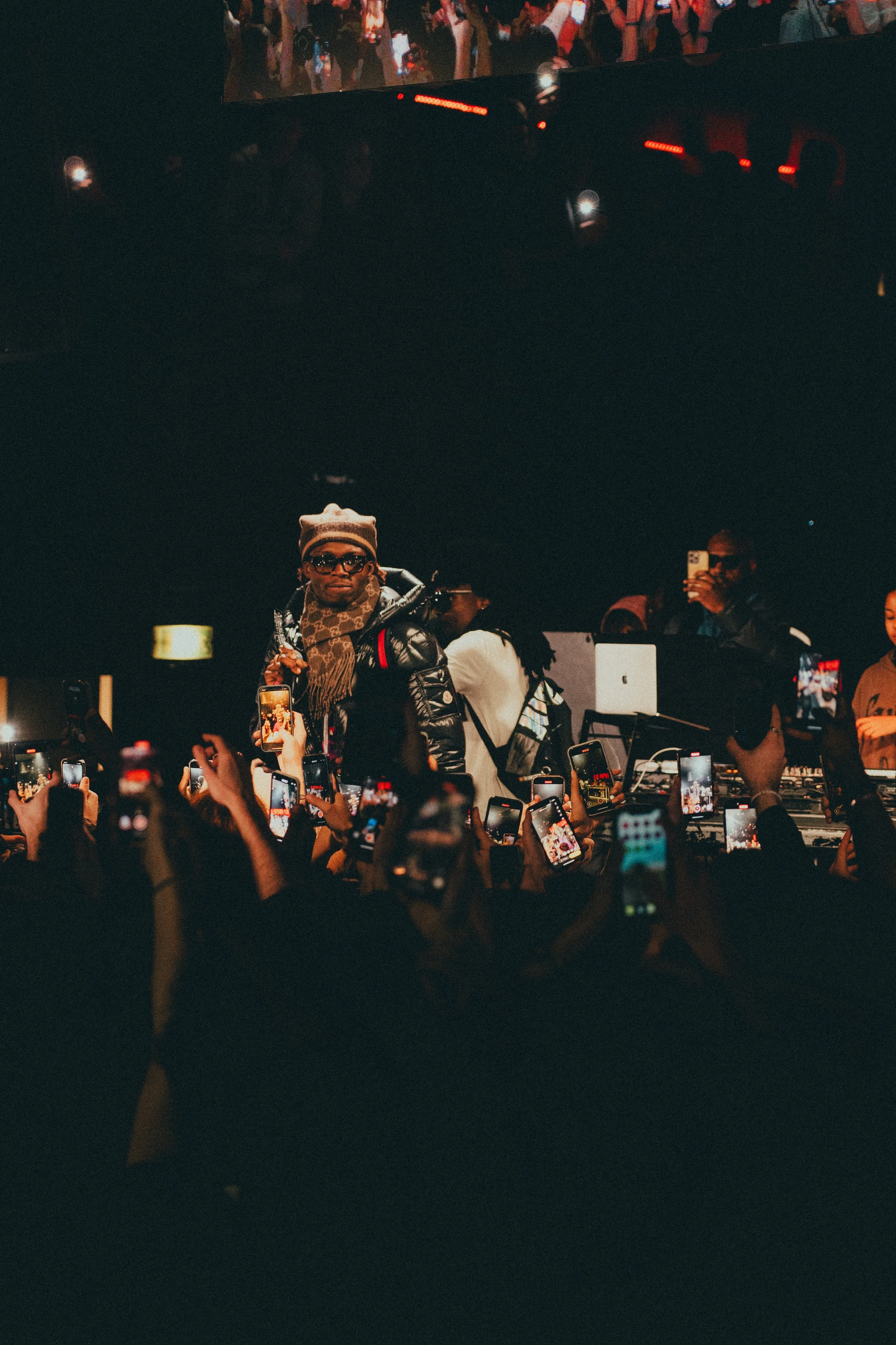 Un DJ sur scène entouré de nombreux spectateurs tenant des téléphones portables, dans une ambiance de concert ou de club nocturne.