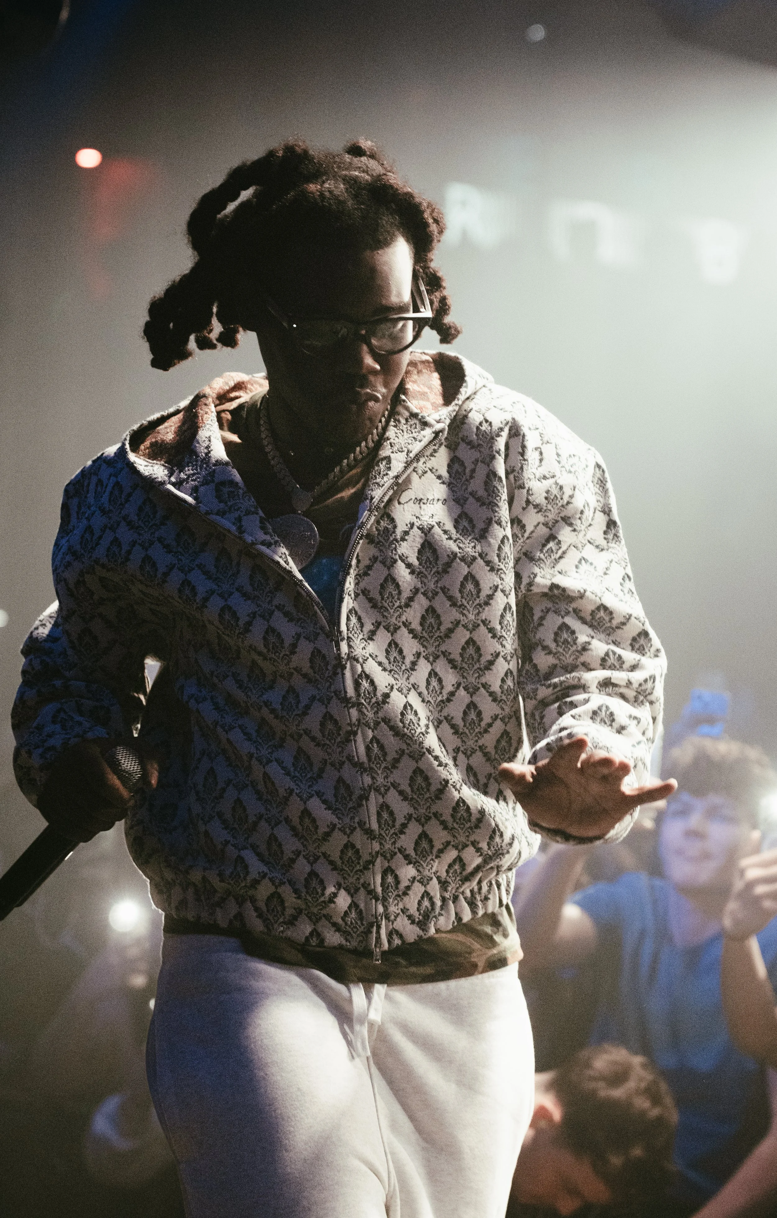 Un homme avec des dreadlocks portant des lunettes, une veste à motif et des sweatpants blancs, tenant un microphone en scène, dans une ambiance sombre avec des personnes qui prennent des photos en arrière-plan.