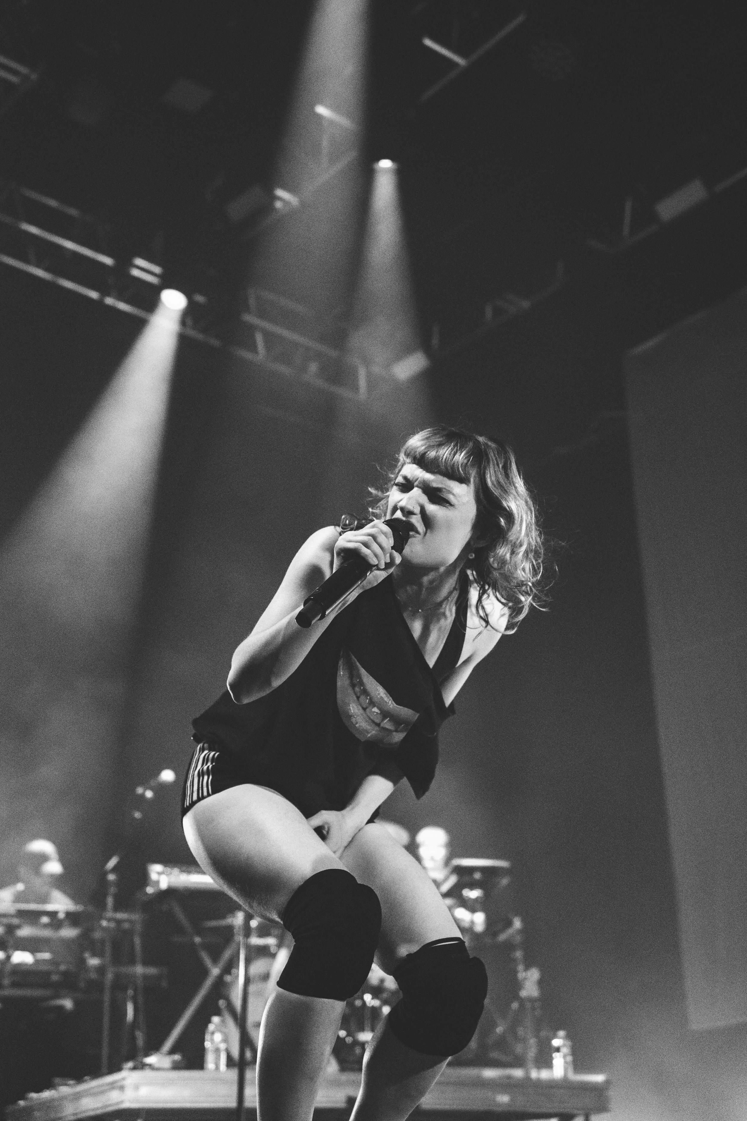 Une femme chante en scène avec des lumières de scène et un microphone dans une atmosphère sombre et concert. Elle porte un débardeur noir et des chaussettes hautes.