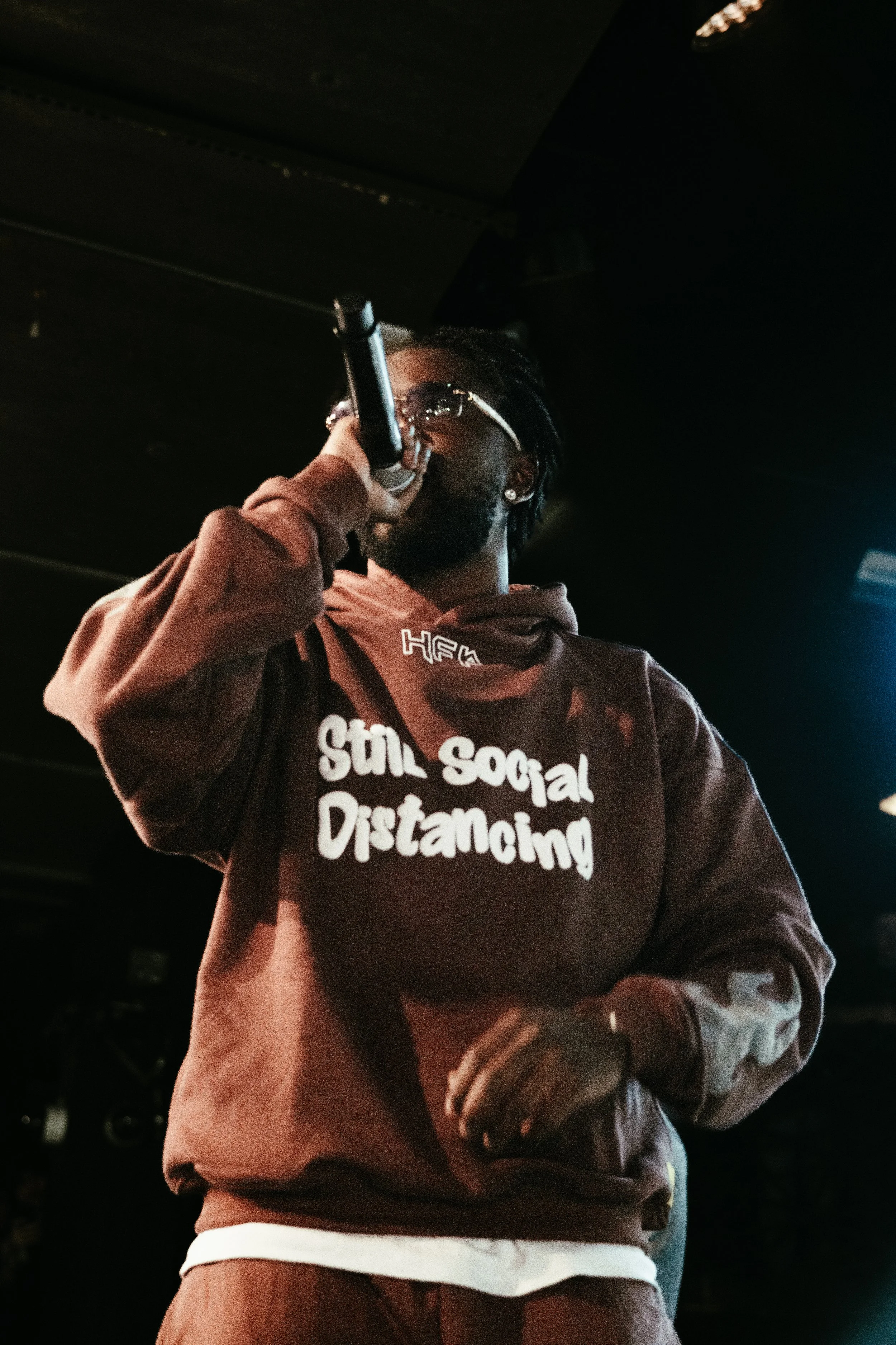 Homme portant une veste marron avec l'inscription « Still Social Distancing » en blanc, tenant un microphone, dans une ambiance sombre.
