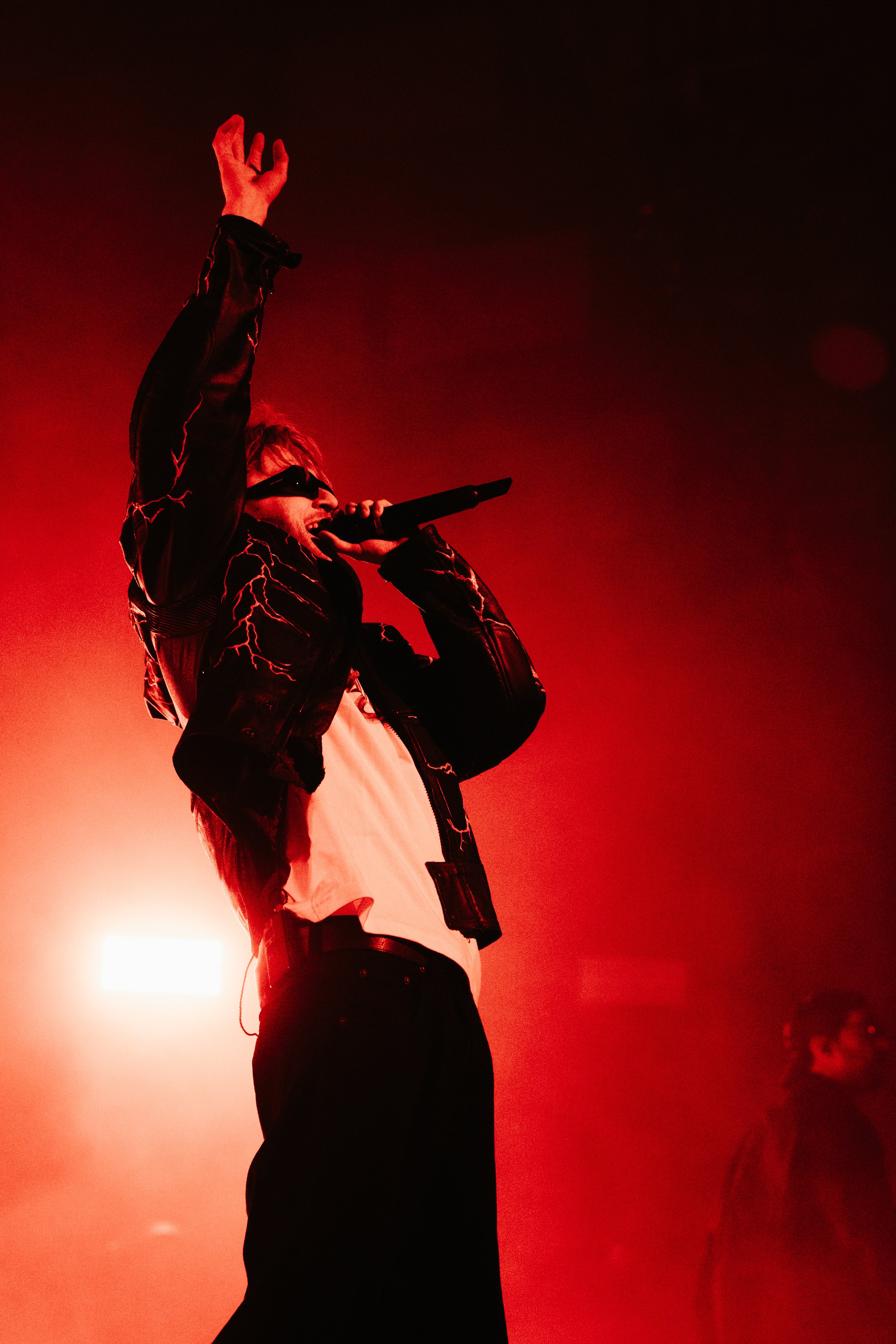 Un chanteur portant des lunettes de soleil et un blouson à motifs de lightning, tenant un microphone, en train de performer sur scène avec un éclairage rouge.