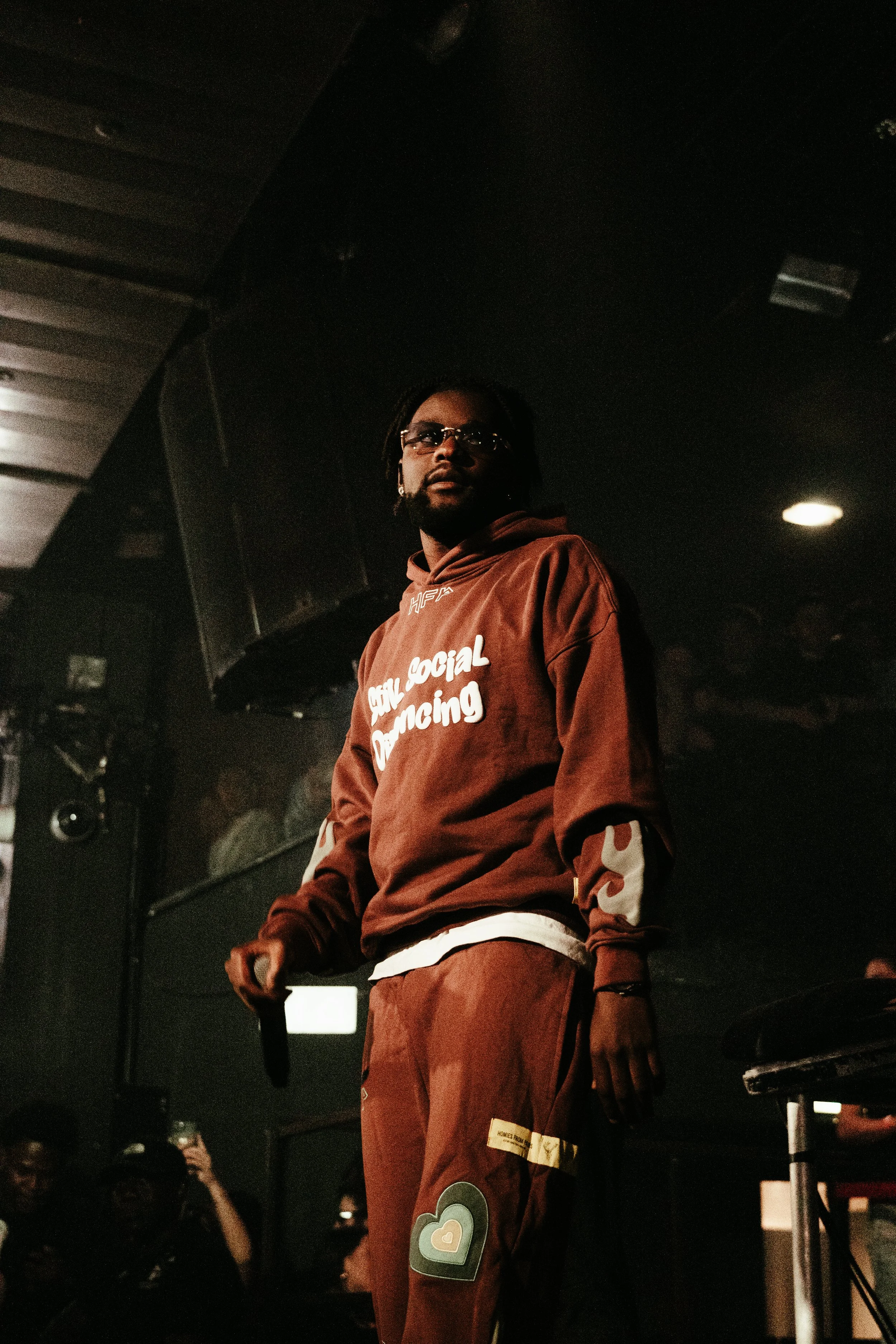 Un homme noir porte un sweat à capuche marron avec des écritures blanches et un pantalon assorti, tenant un microphone dans une main, dans un lieu sombre, probablement lors d'une performance ou d'un événement.