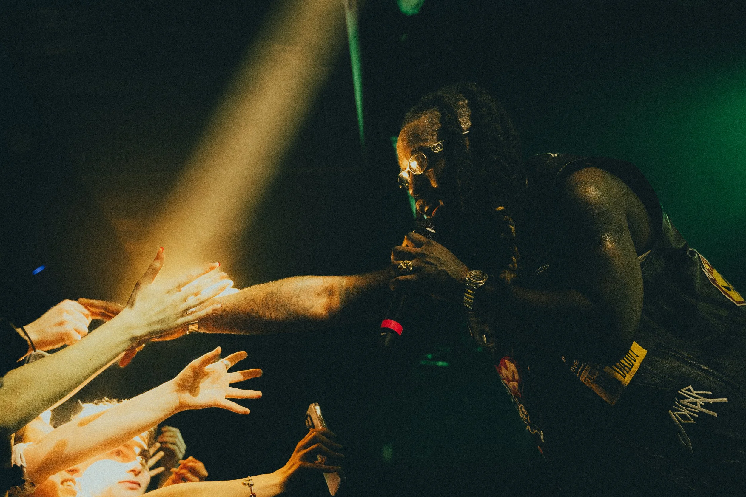 Un chanteur de reggae en performance sur scène, portant des lunettes rondes, une veste en cuir, tenant un micro, en train de chanter et d'interagir avec le public, dans un éclairage sombre avec des lumières vertes.